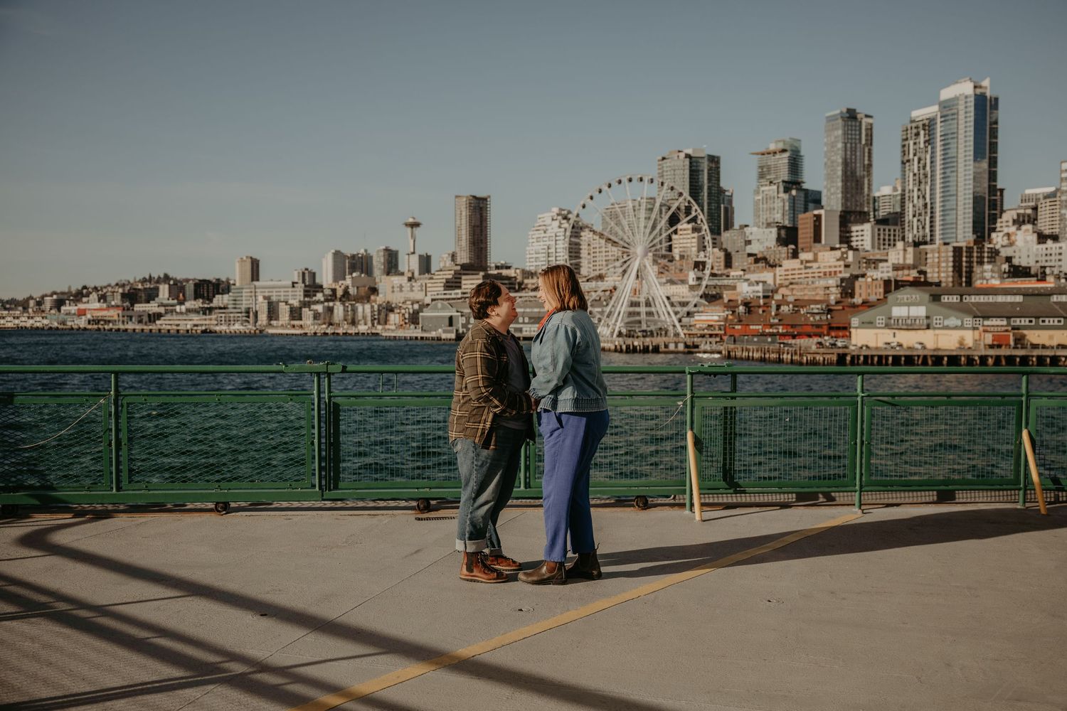 Couple holding hands on Seattle waterfront with Space Needle, Ferris wheel, and city skyline in background.