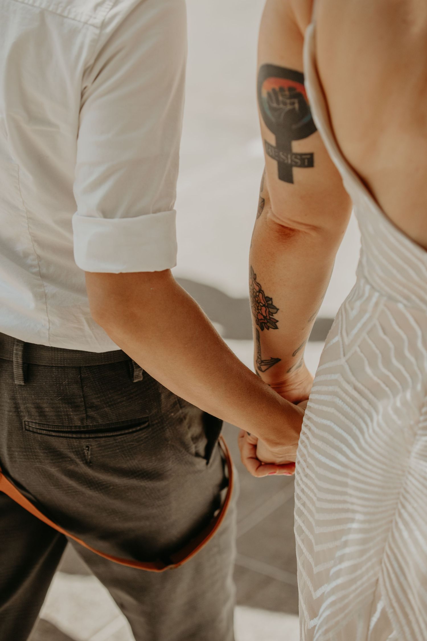 Close-up of a couple holding hands, woman wearing a white dress with tattoos, man in white shirt and gray pants.