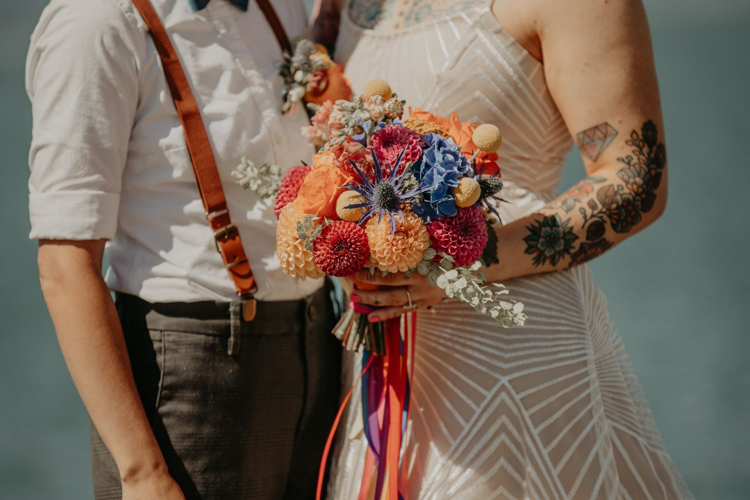 A couple holds a vibrant wedding bouquet with red, blue and orange flowers tied with colorful ribbons.