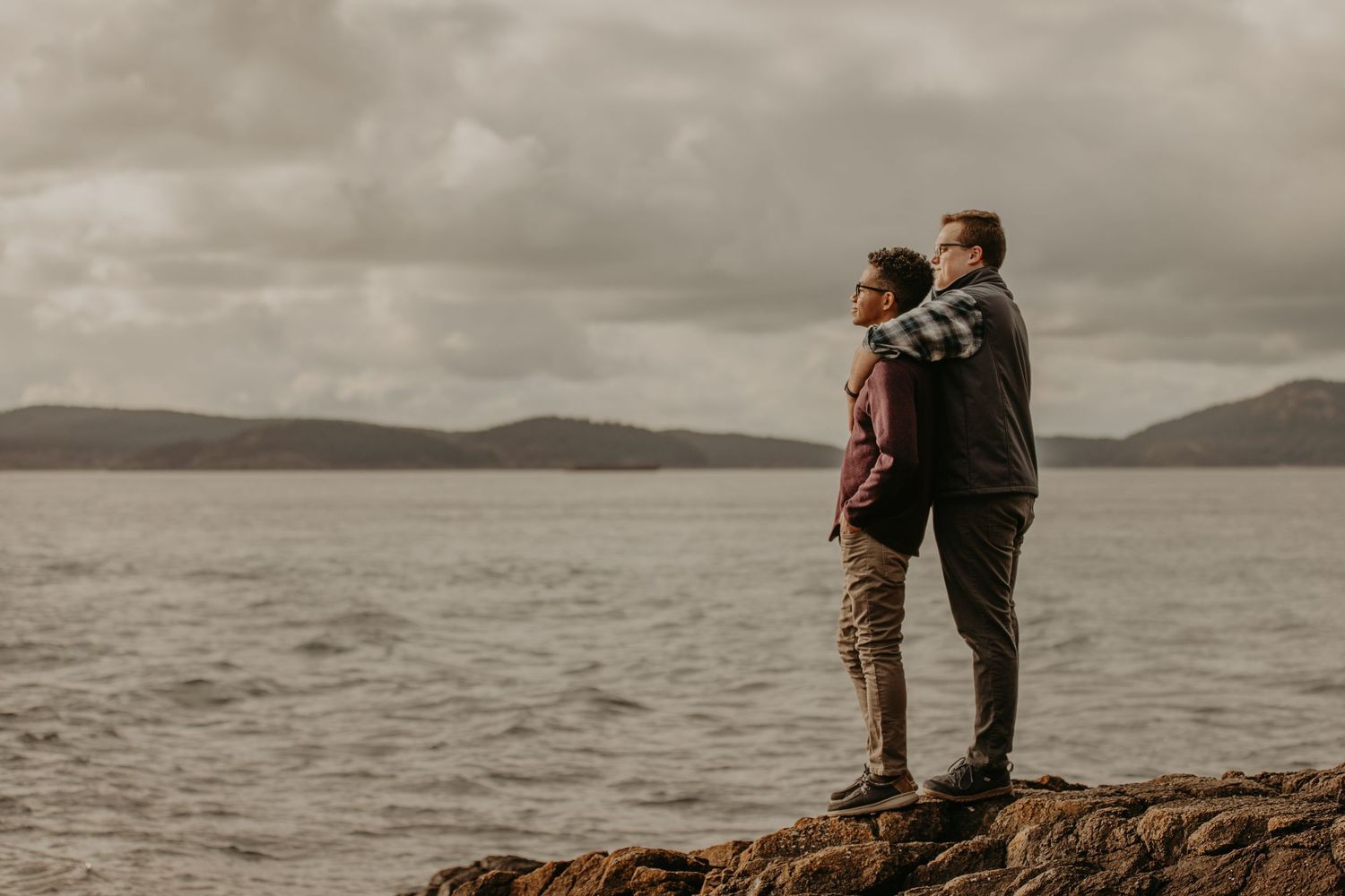 Two people stand on a rocky cliff overlooking a moody lake with mountains in the distance under cloudy skies.