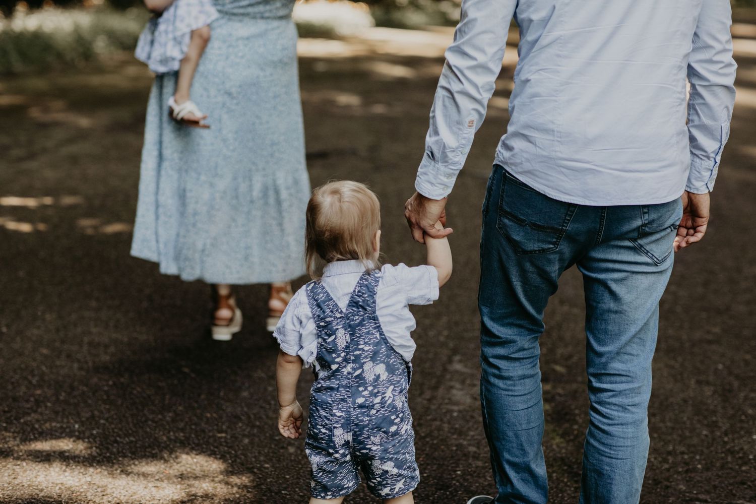 A toddler in floral overalls holds hands with their parents while walking along a shaded path outdoors.