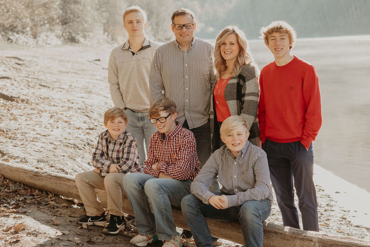 A family of seven wearing casual clothing poses together on a wooden log at a scenic beach on an overcast day.