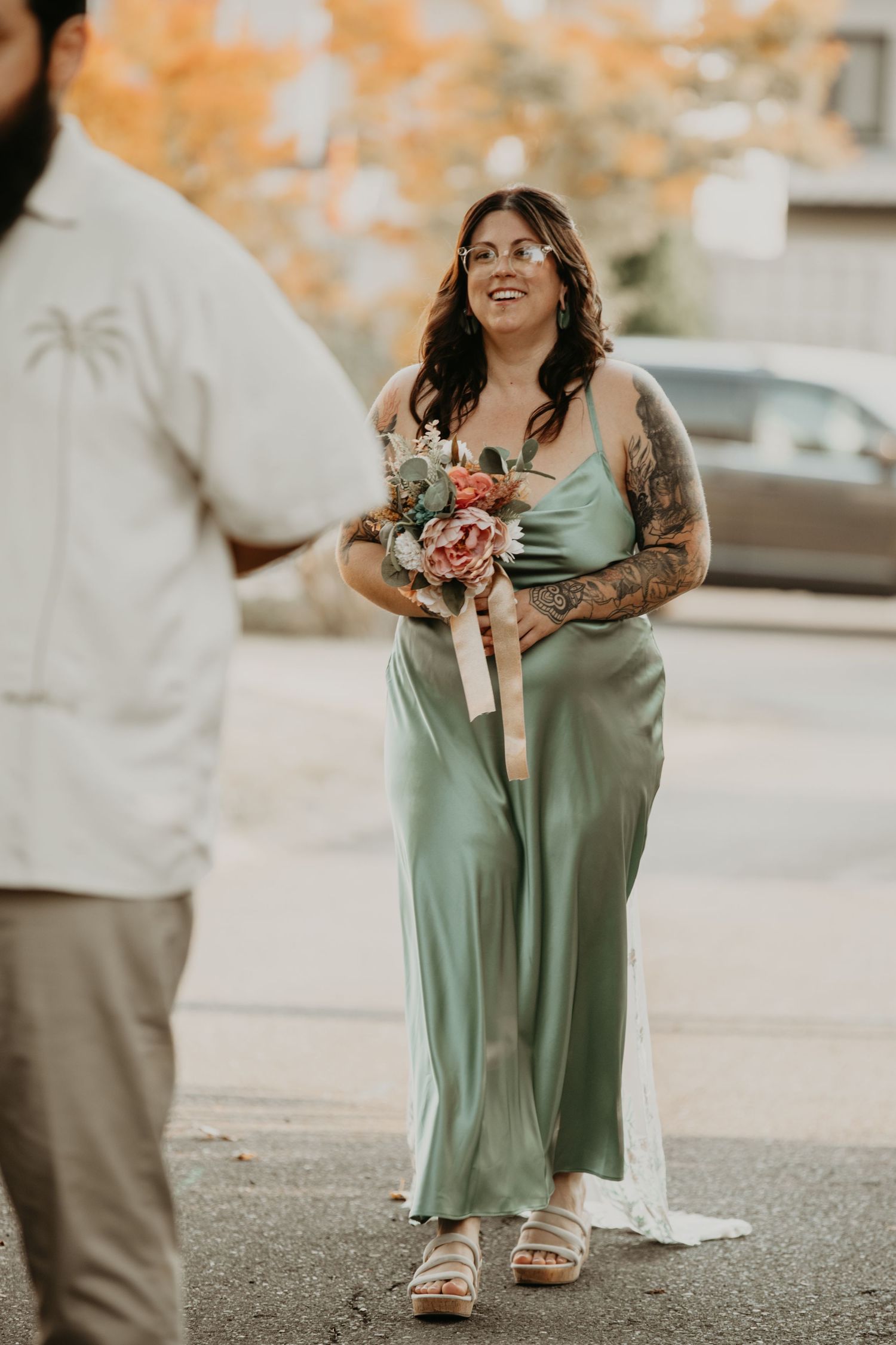 Person in flowing sage green dress holding floral bouquet stands outdoors on autumn day wearing sandals.