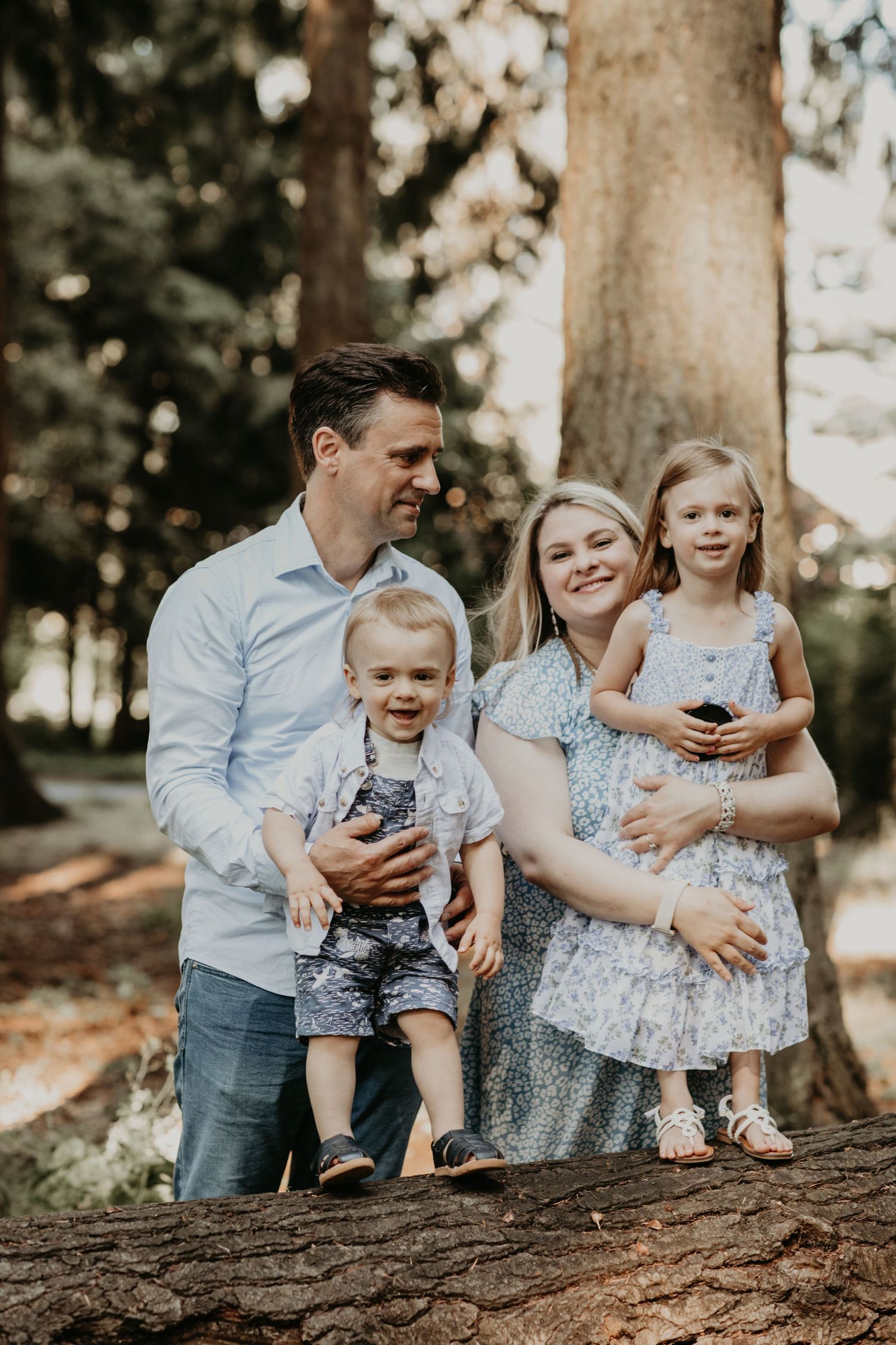 Family portrait in a forest setting standing on a fallen tree trunk with soft natural lighting and earthy tones.