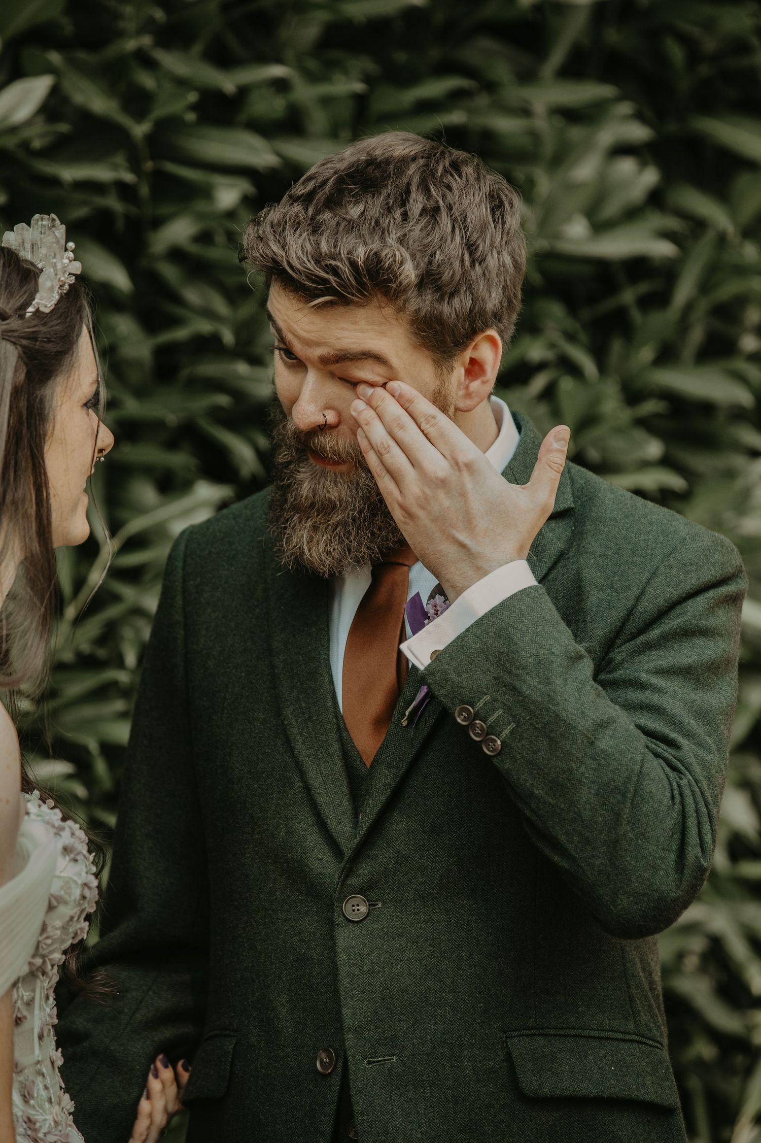 Emotional first look reaction between newlyweds in a forest setting, with groom in dark green suit covering his face.