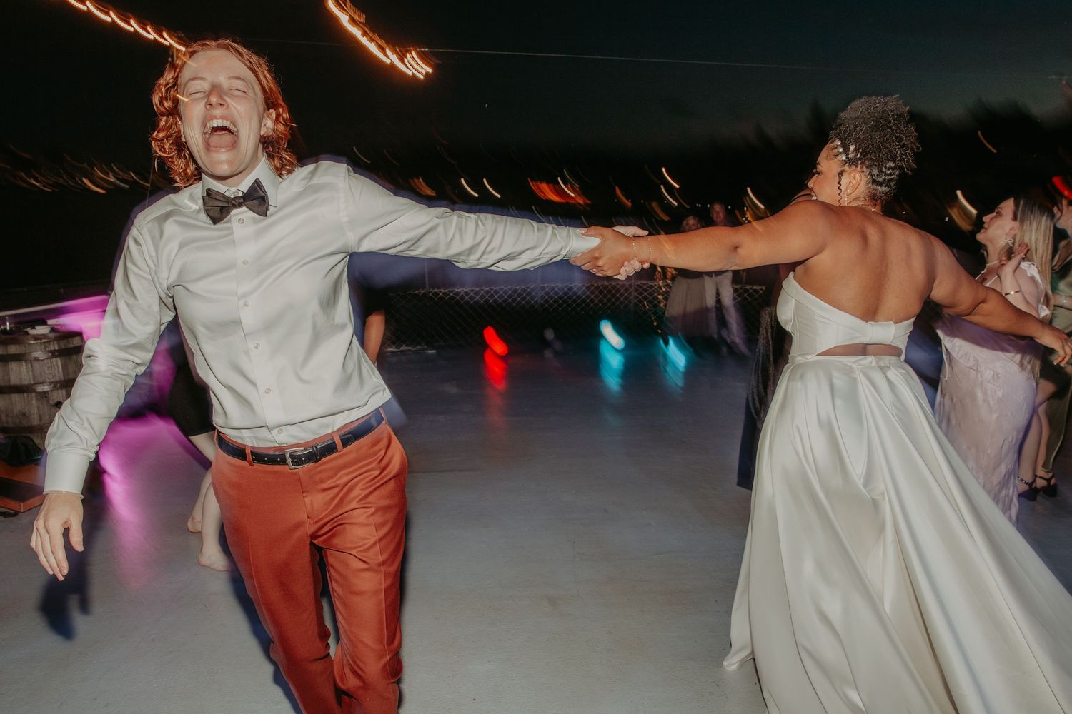 Wedding party dancing energetically on a dark dance floor with colorful lighting.