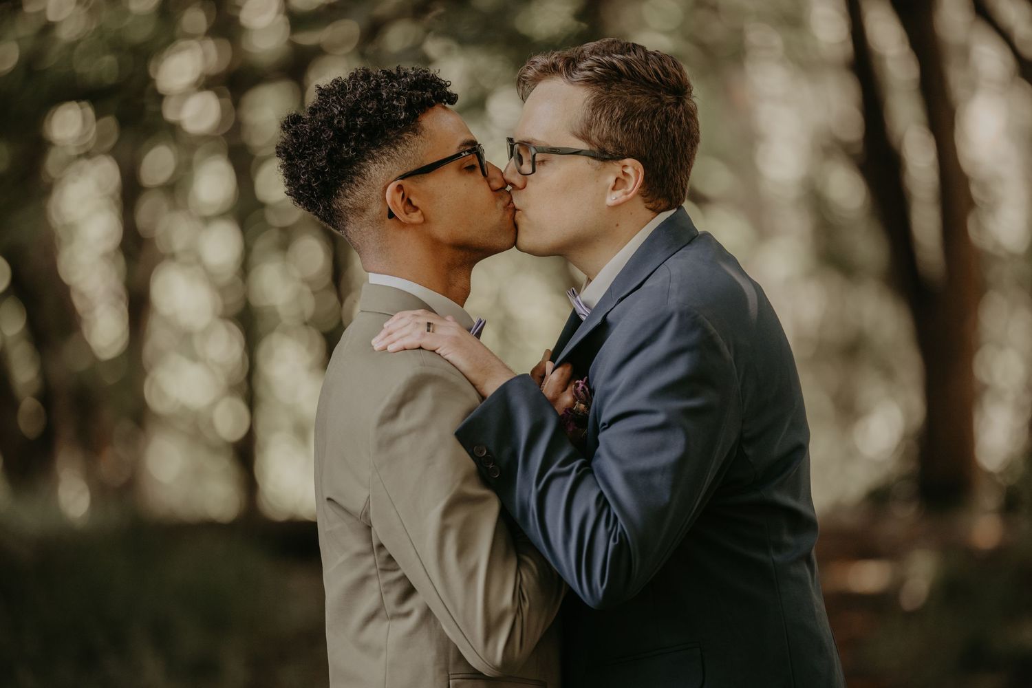 A romantic couple sharing an intimate kiss outdoors in a wooded setting with soft natural lighting.