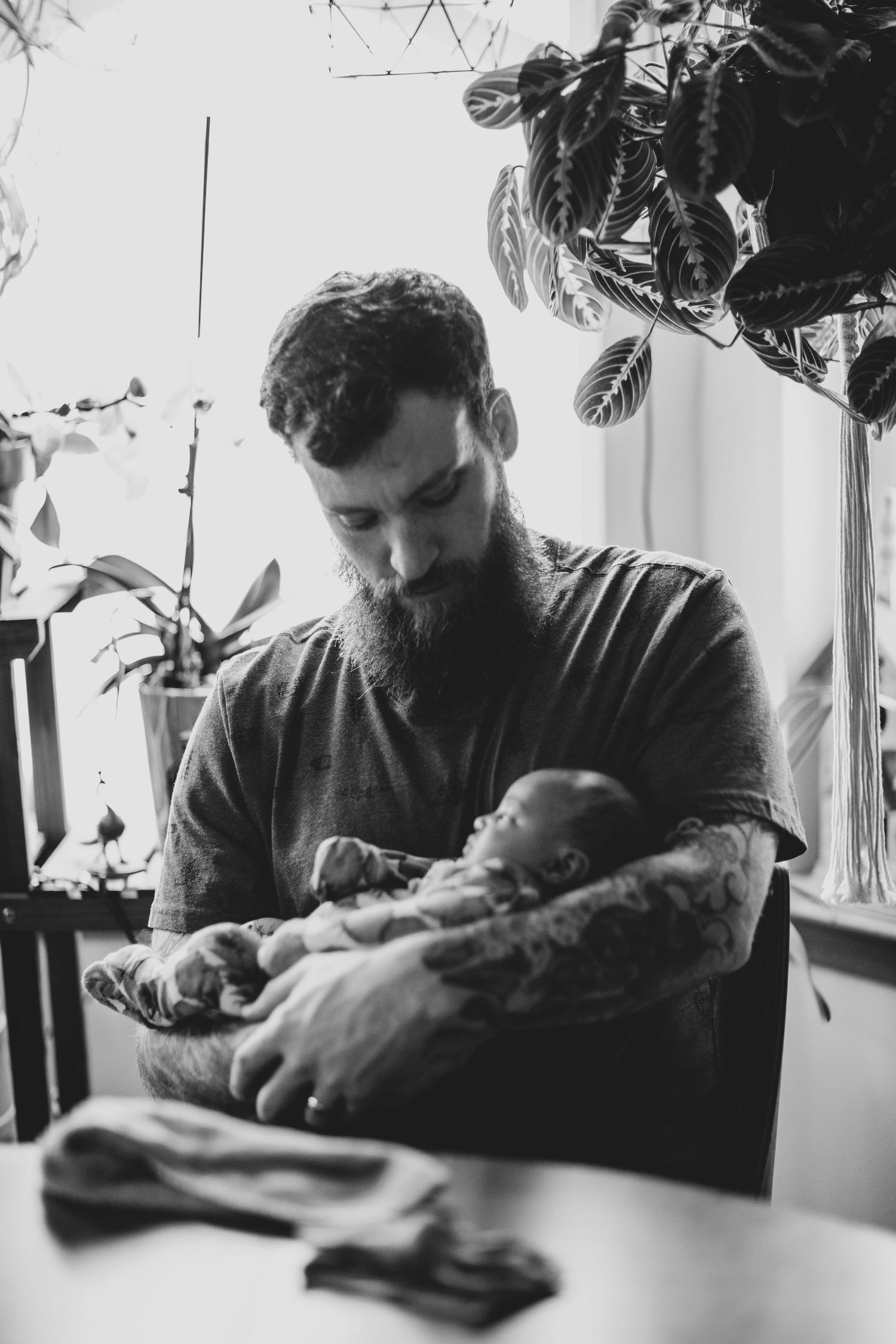 Black and white photo of a bearded man in dark shirt holding sleeping newborn near houseplants by window.
