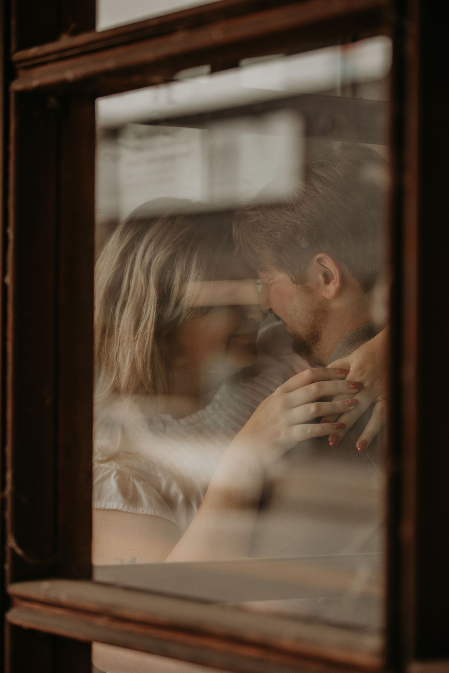 An intimate moment seen through a window shows a couple embracing in soft, dreamy lighting.
