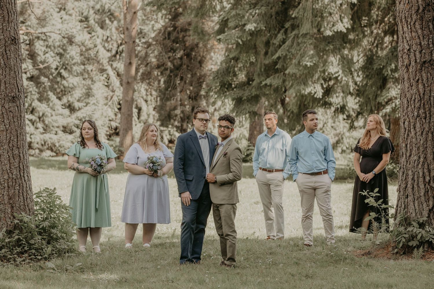 Wedding party stands together outdoors in a casual backyard ceremony setting surrounded by tall trees.