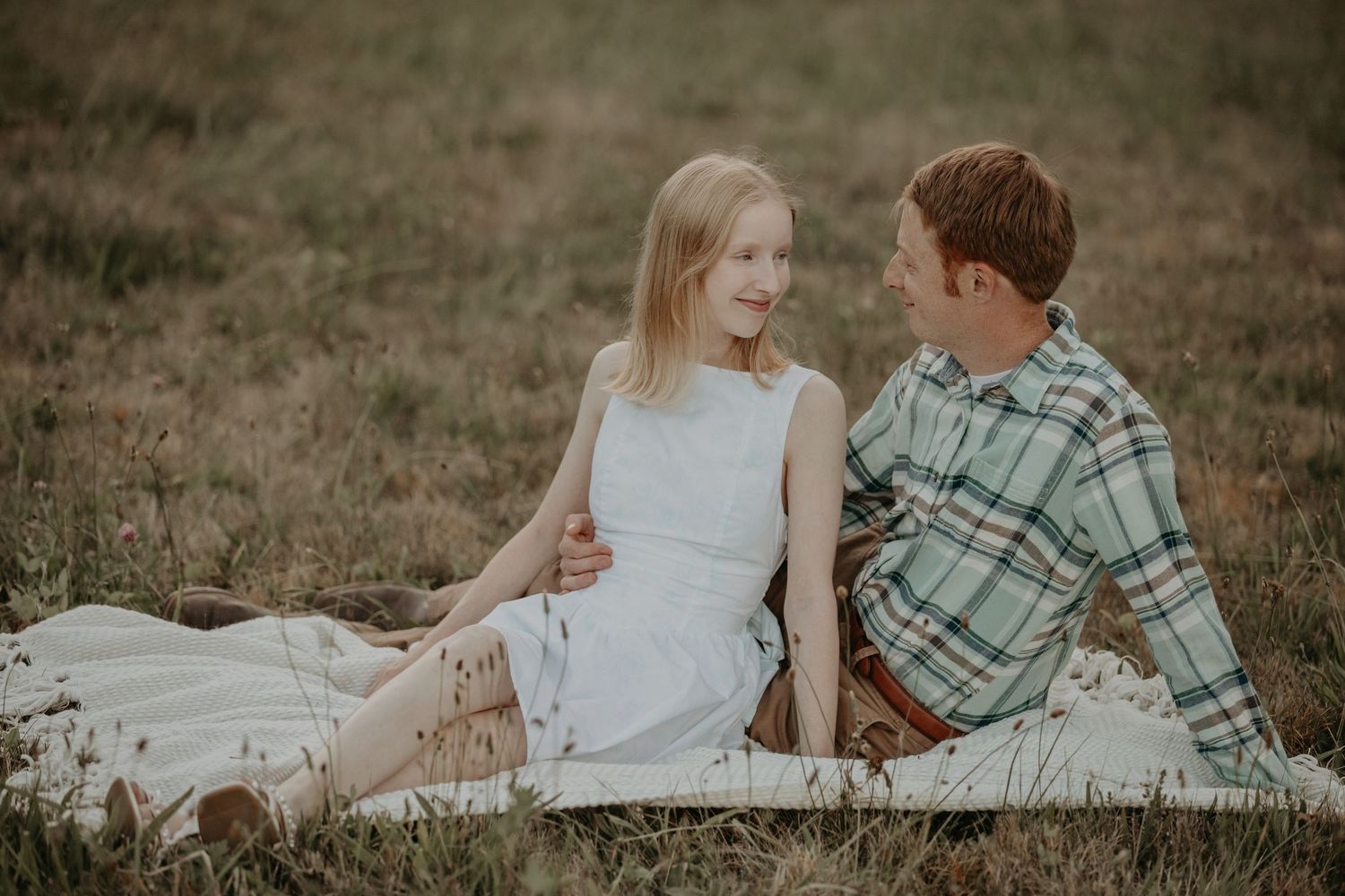 Intimate couple sitting together on a blanket in a field during golden hour in desaturated tones.