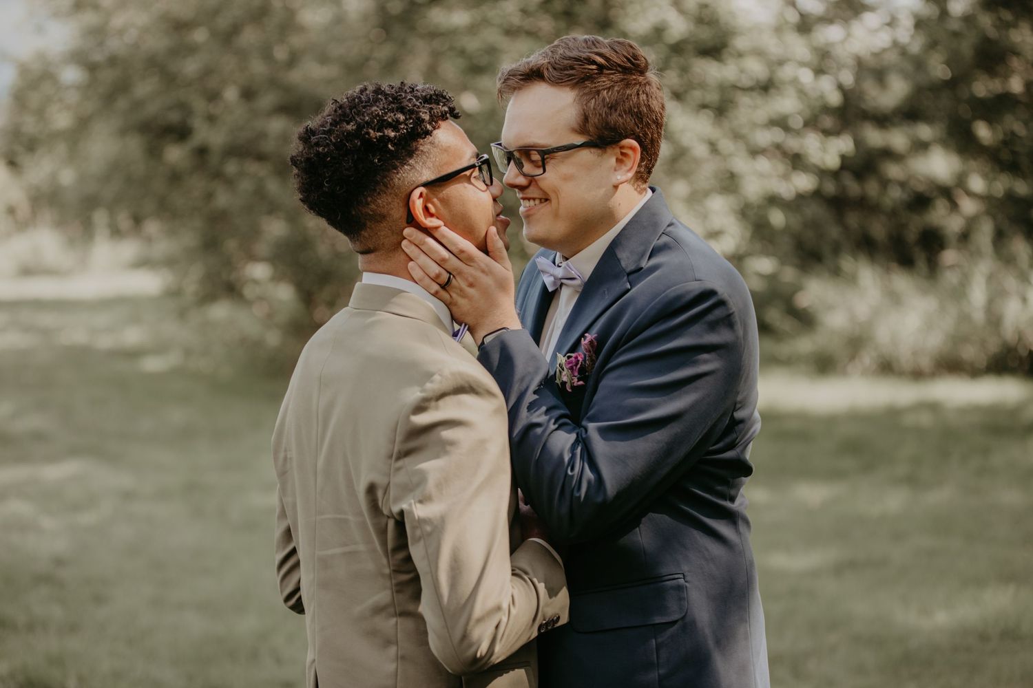 Two people in suits share an intimate moment on a grassy lawn with trees in the background.