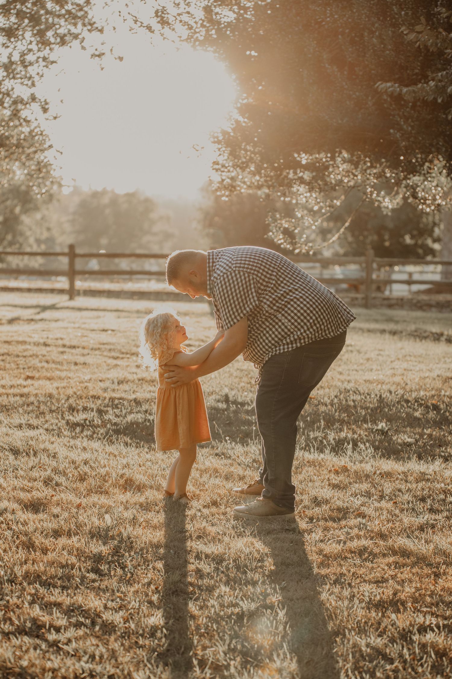 A tender moment between parent and child in a sunlit field with wooden fencing in the background.