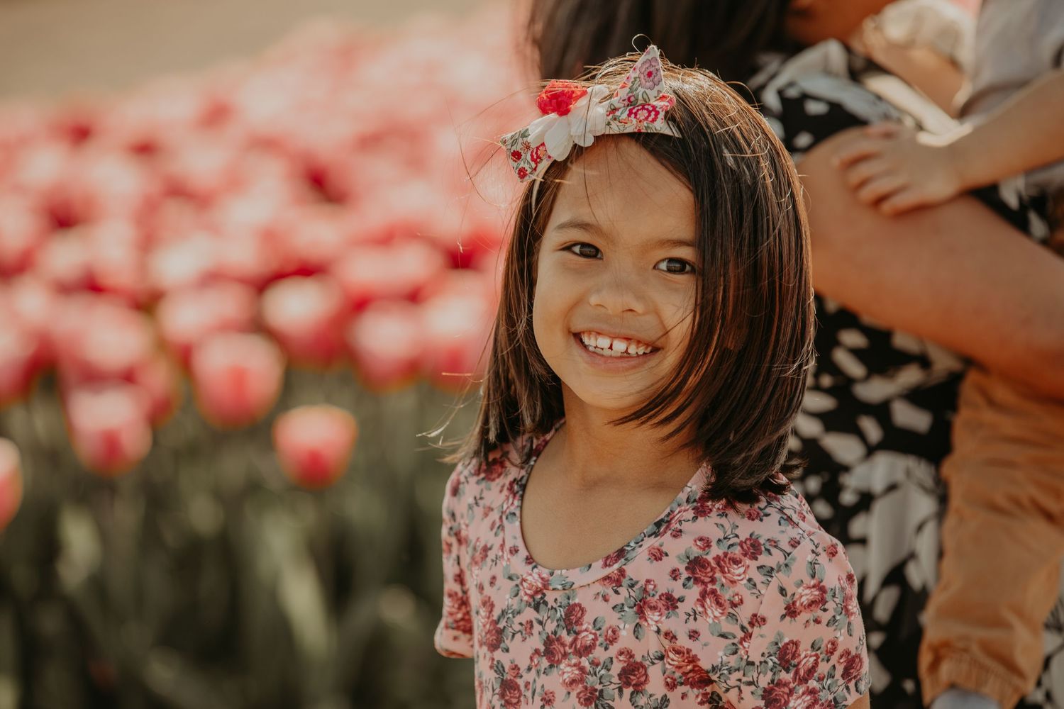 Child smiling in a pretty floral dress standing in front of a field of pink tulip flowers.