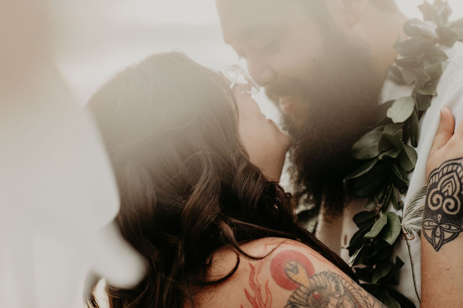 Romantic couple sharing a kiss in soft sunlight with white flowers and visible arm tattoos.