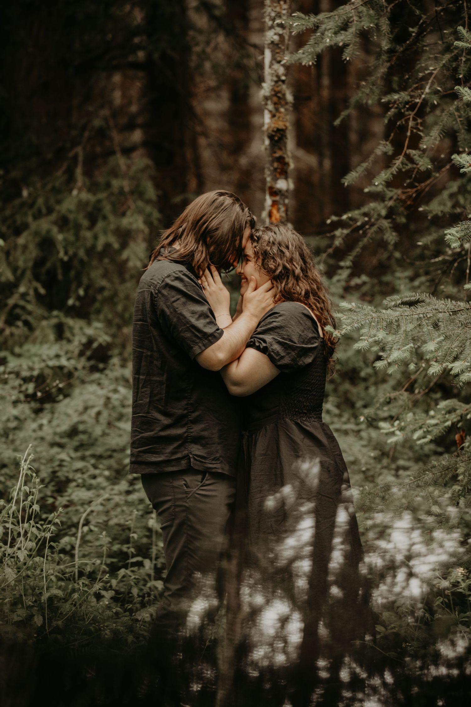 A romantic couple shares an intimate moment in a moody forest setting with soft natural lighting.