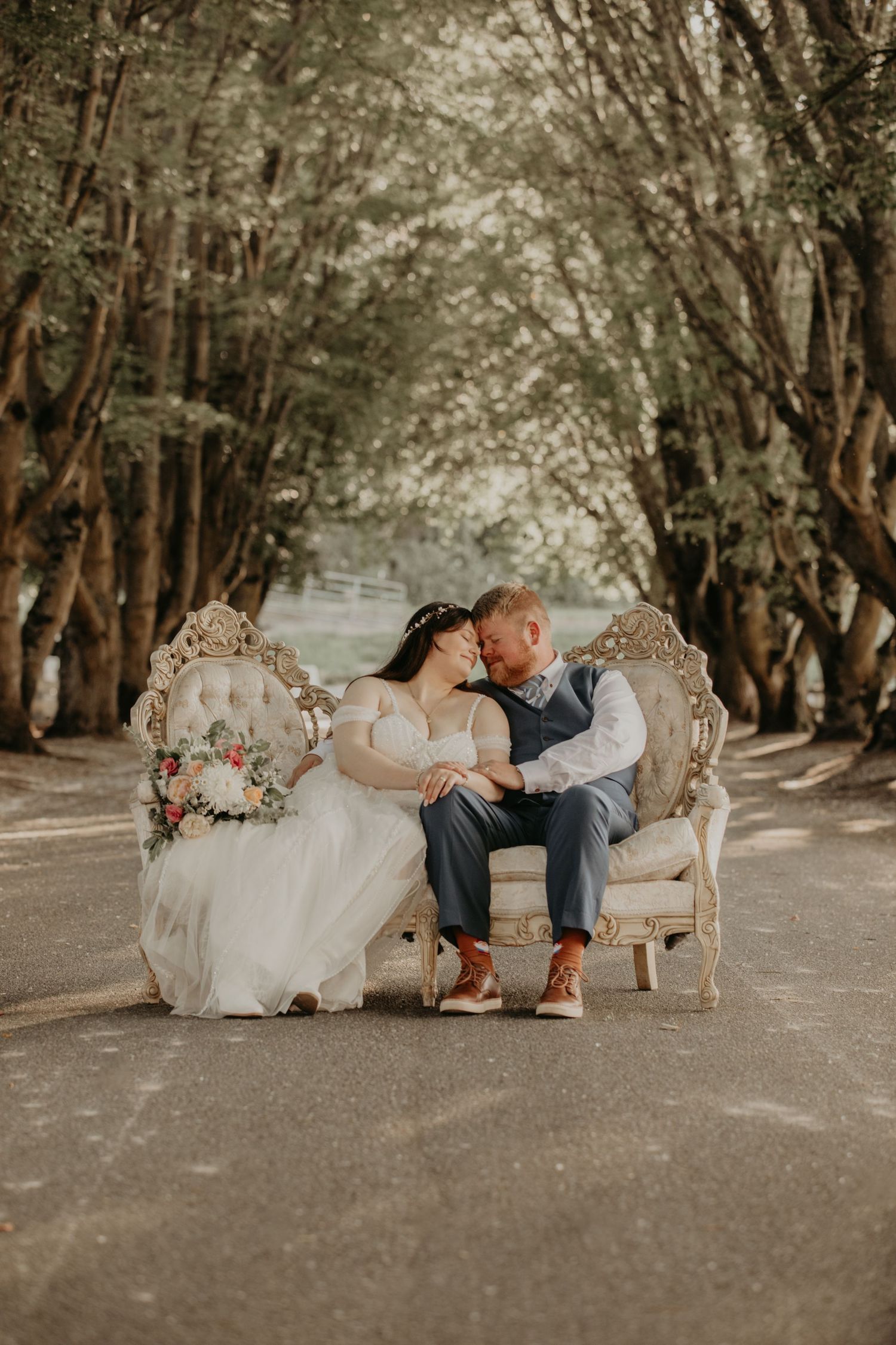 Beautiful wedding couple shares intimate moments on vintage cream sofa set along tree-lined path during outdoor photoshoot.
