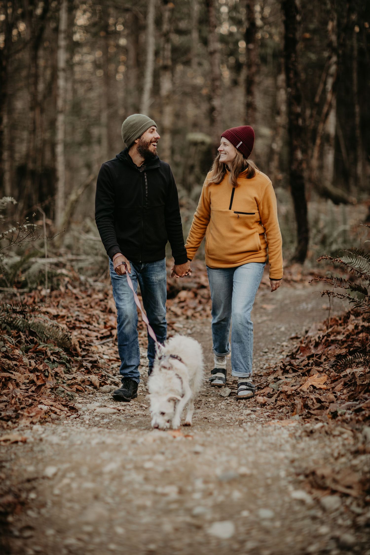 Two people in warm layers walk their fluffy white dog on a forest trail covered in fallen autumn leaves.