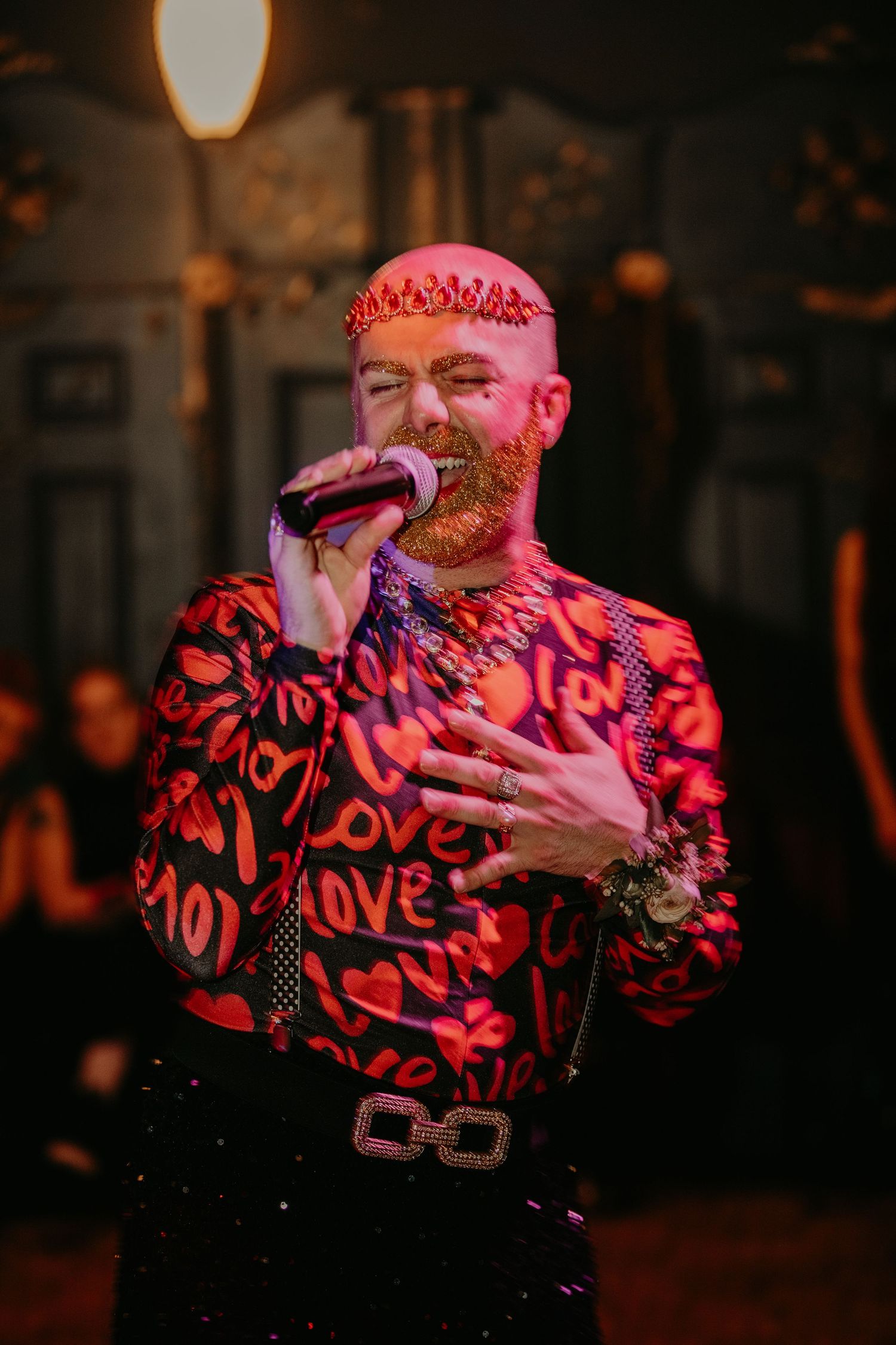 A performer in a red patterned shirt with a sparkly belt sings passionately on stage under warm lighting.
