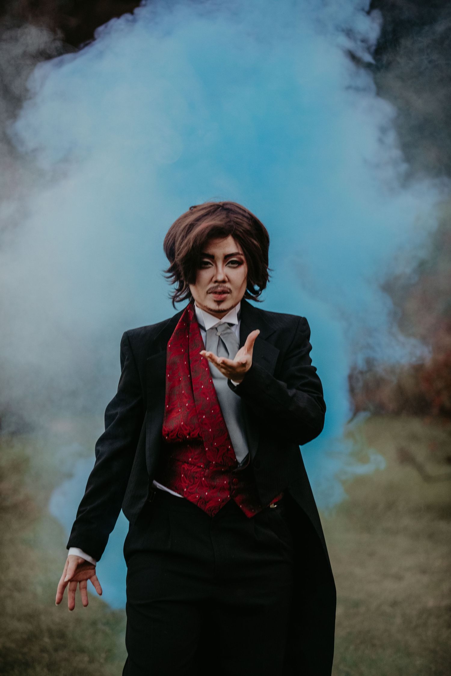 A figure in a black suit and red vest blows magic dust while surrounded by ethereal blue smoke in a mystical scene.