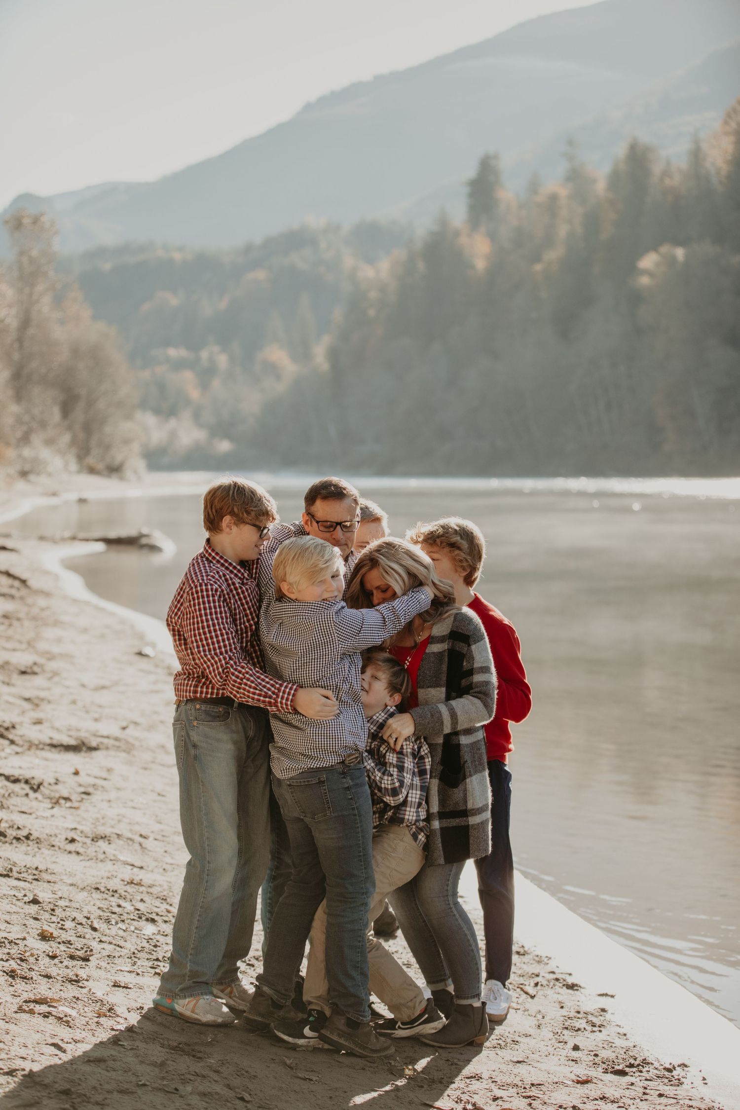 A group stands embracing together on a scenic beach shore with mountains and forest in the background.
