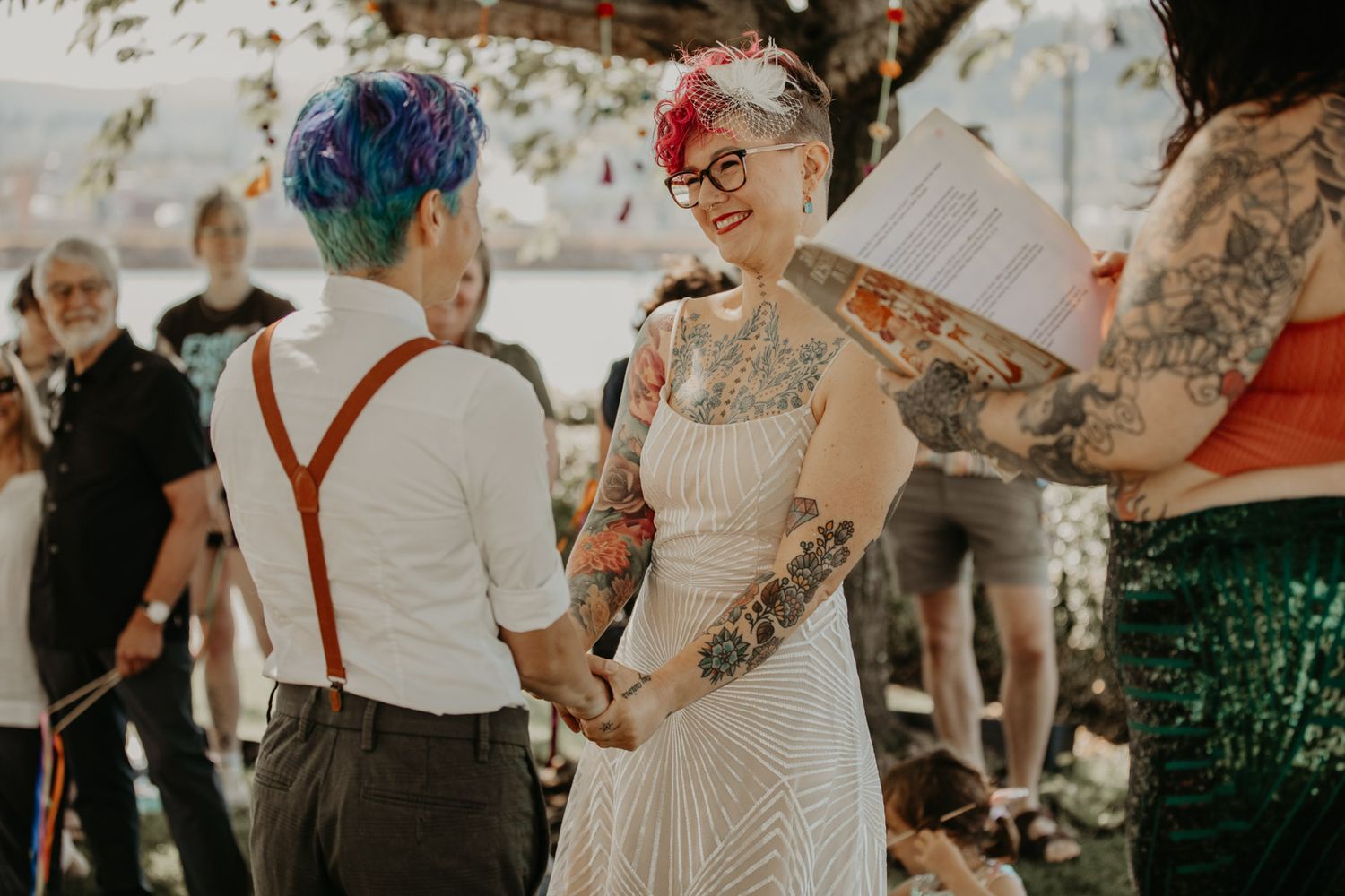 A casual outdoor wedding ceremony with guests in white attire and tattoos visible in natural sunlight.