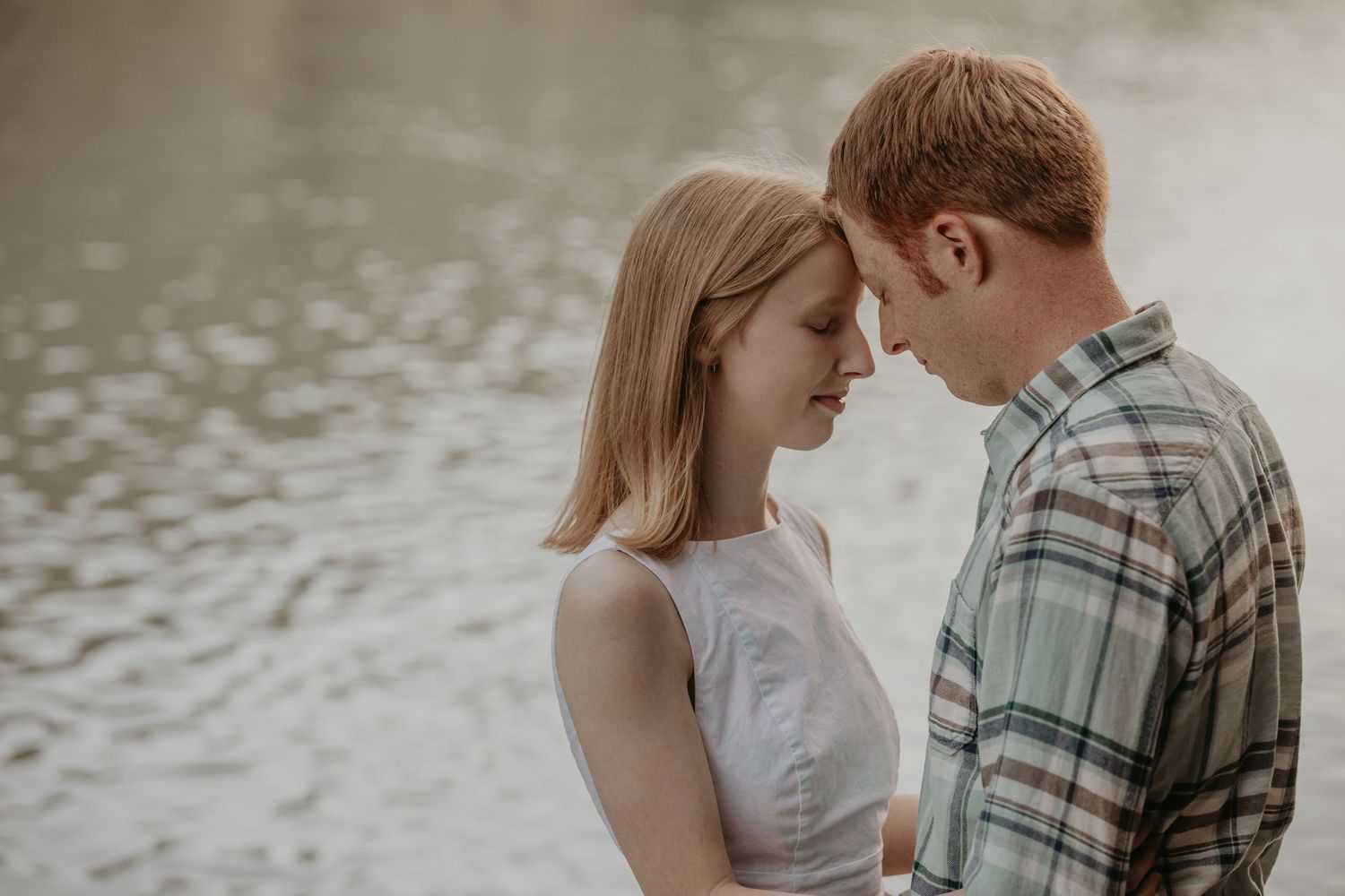 Romantic couple shares tender moments by a serene lake wearing white and plaid during their engagement photoshoot.