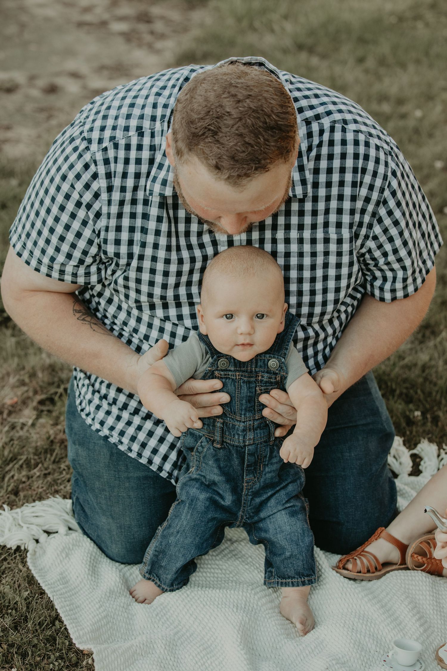 A parent in a checkered shirt holds a toddler wearing denim overalls while sitting on a white blanket outdoors.