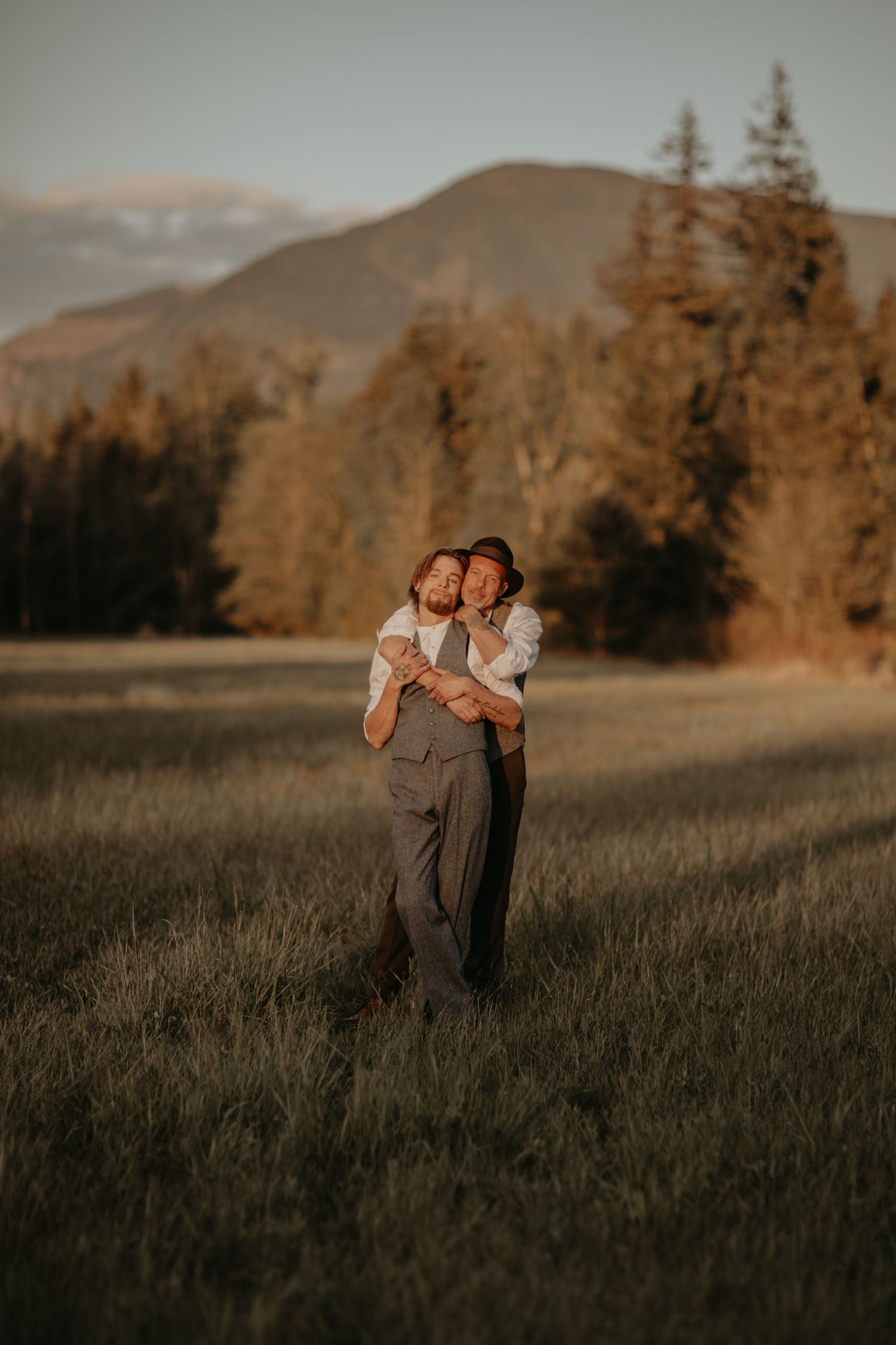 A couple stands intimately in a golden mountain meadow during sunset as soft light illuminates the landscape.