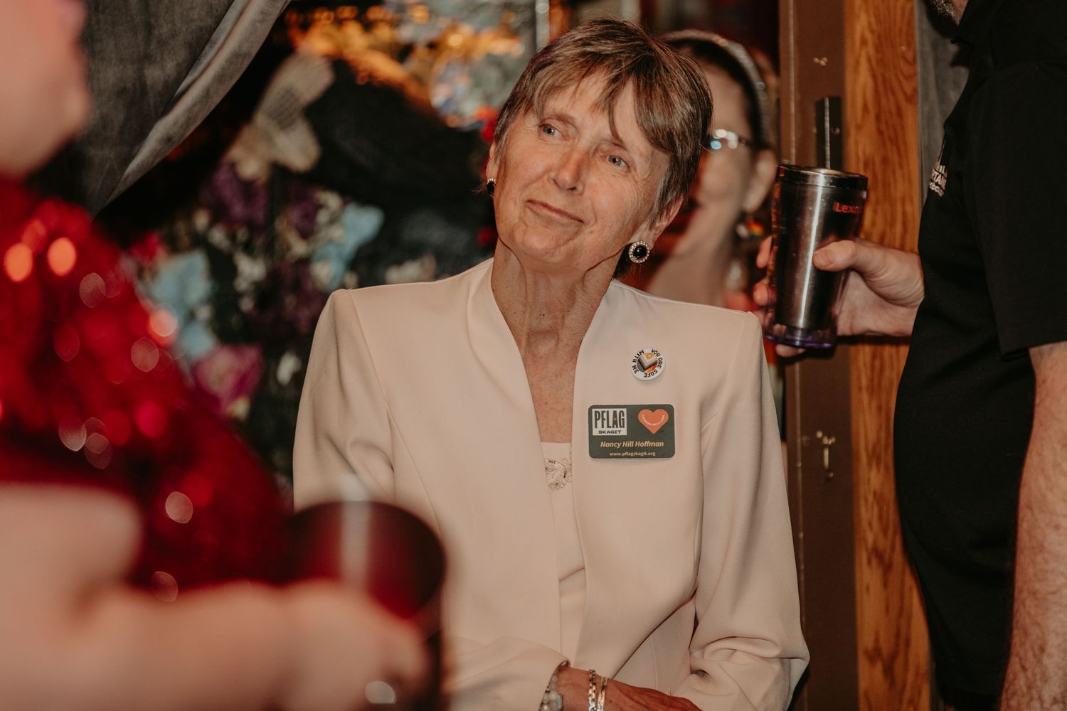A person wearing a beige blazer with a name tag stands next to a wooden pillar at an event.