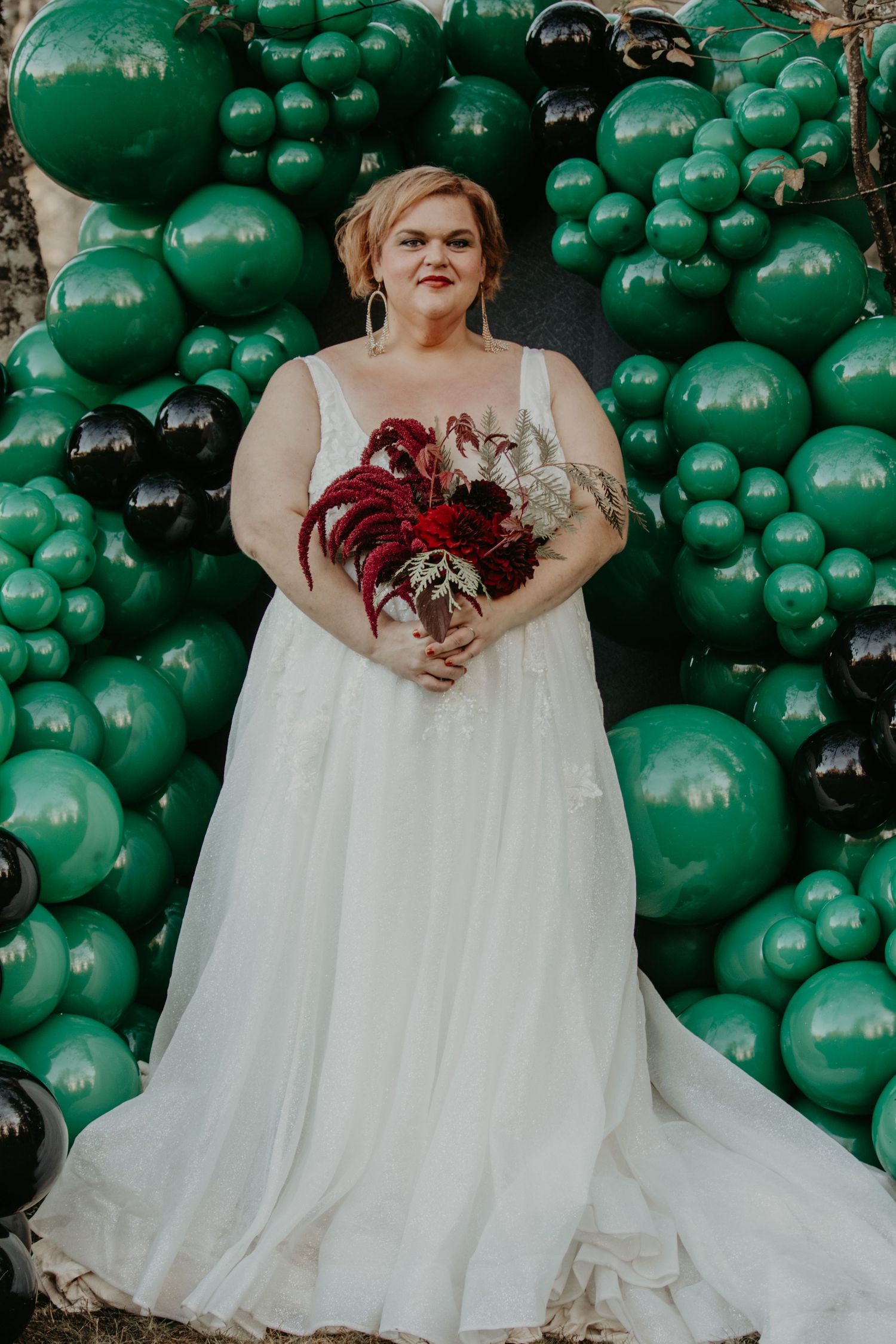 A bride in a flowing white gown holds a dramatic red bouquet against a backdrop of emerald and black balloons.