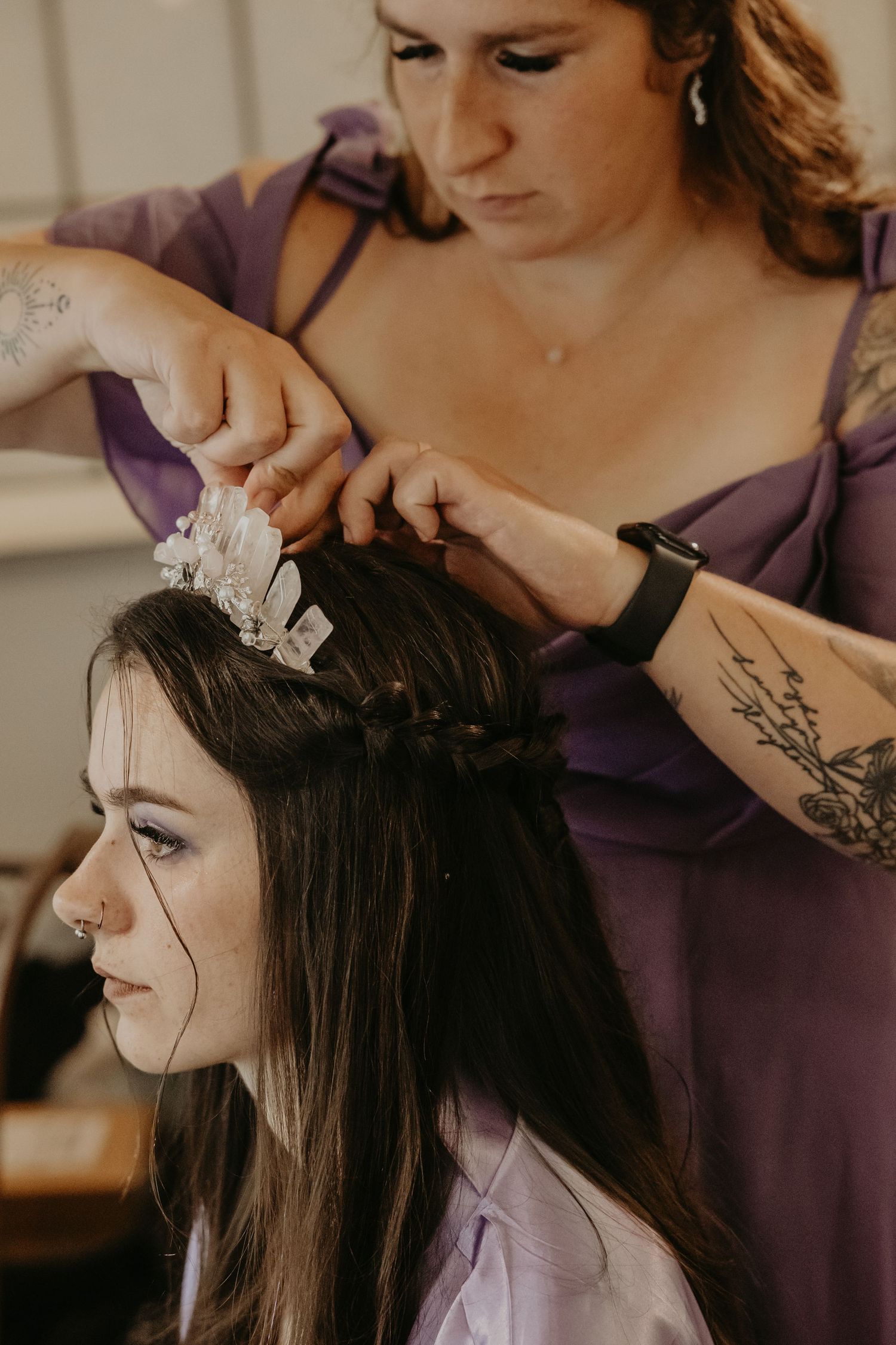 A hairstylist wearing purple applies decorative accessories while styling a bride's hair.