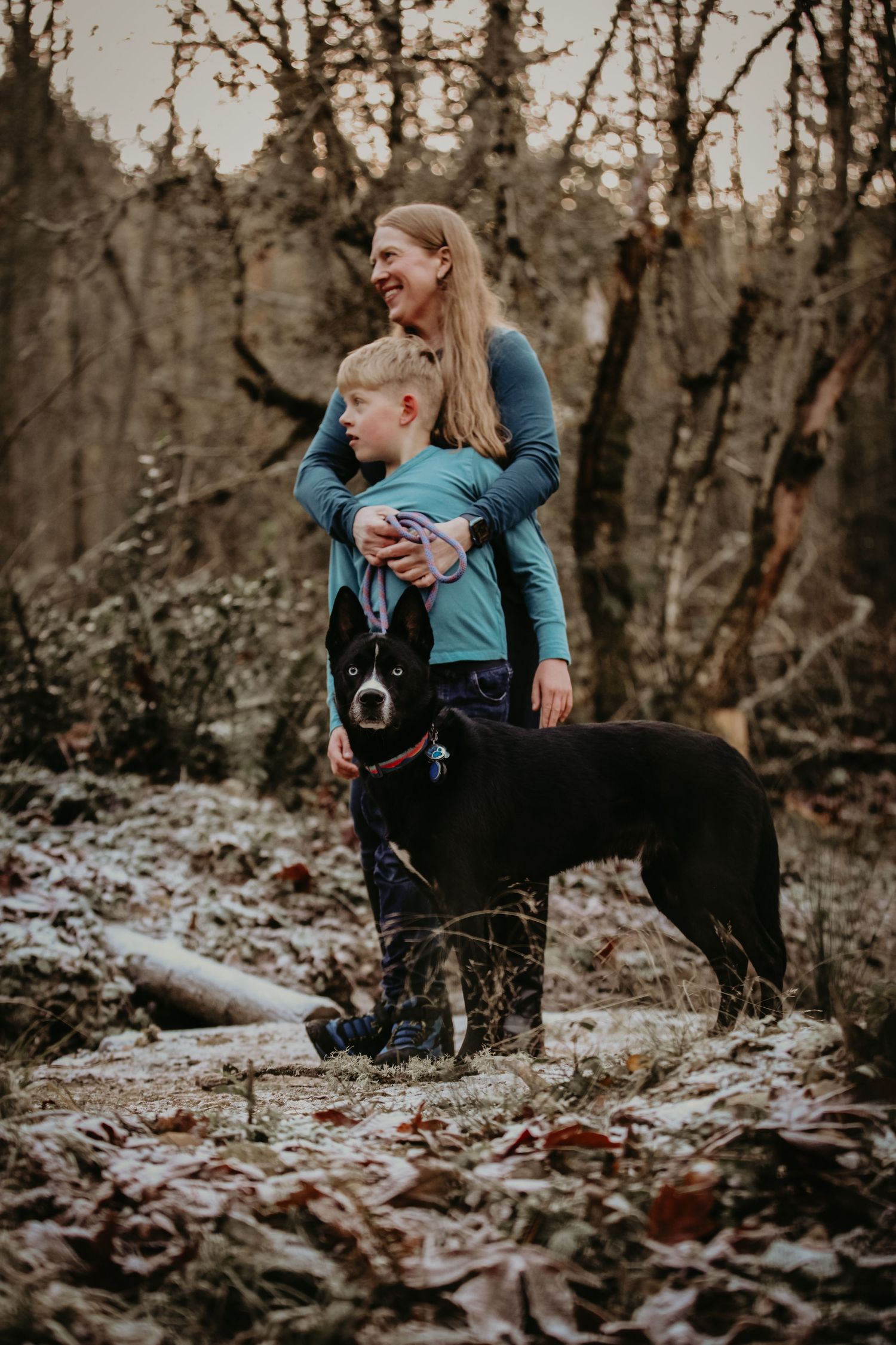 Two people in blue jackets explore autumn woods with their black dog on fallen leaves during an outdoor hike.
