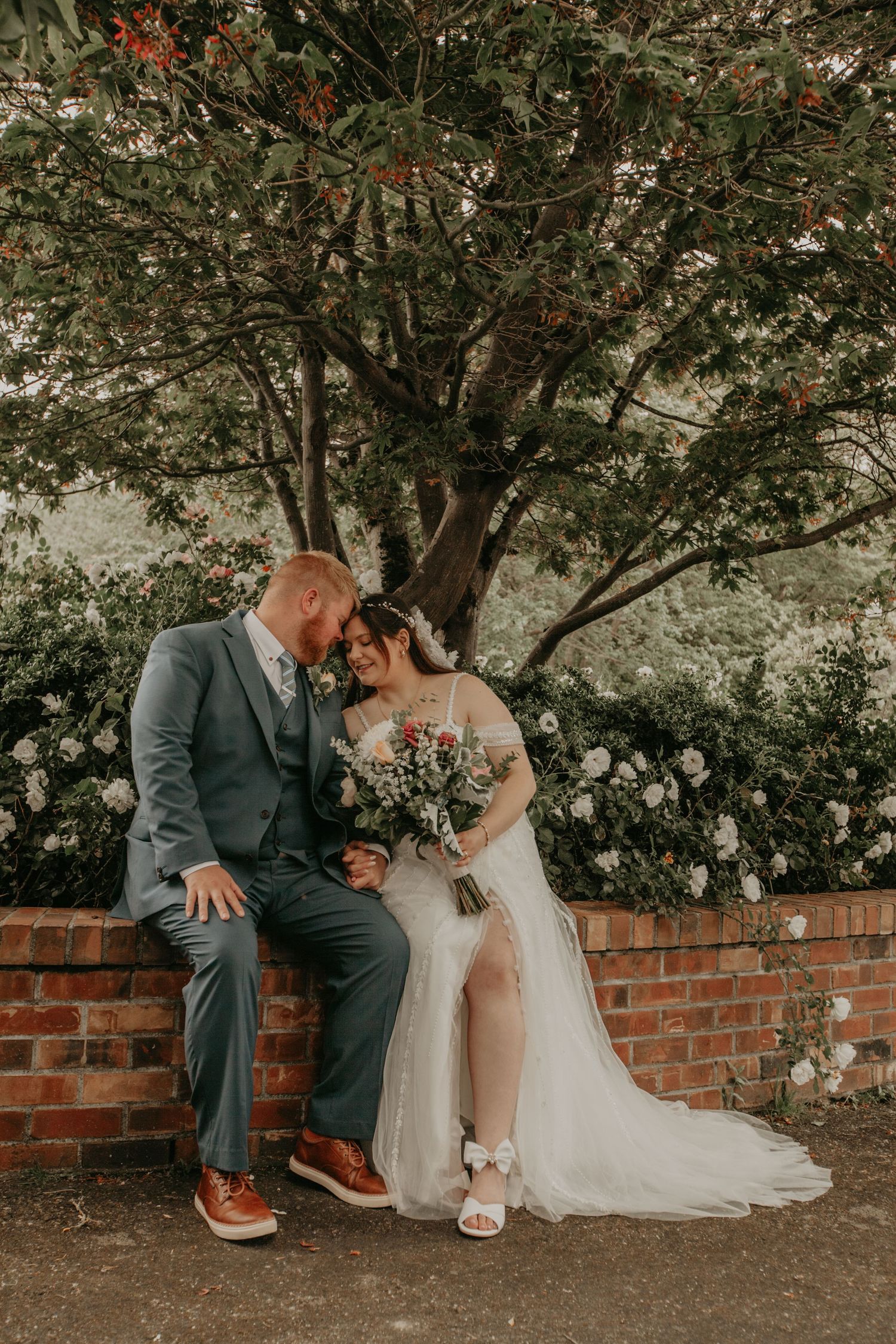 A series of four intimate wedding photos showing a couple sitting together on a brick wall amidst green trees and foliage.