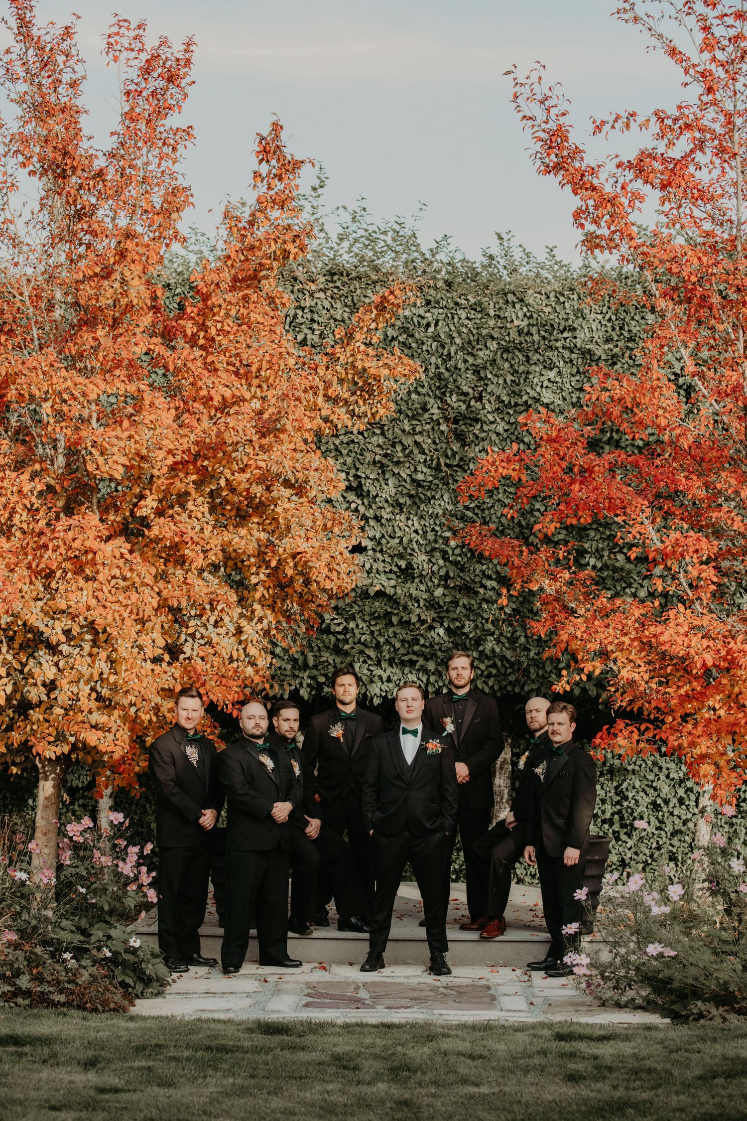 A wedding party stands under vibrant autumn trees with bright orange foliage against a natural outdoor backdrop.