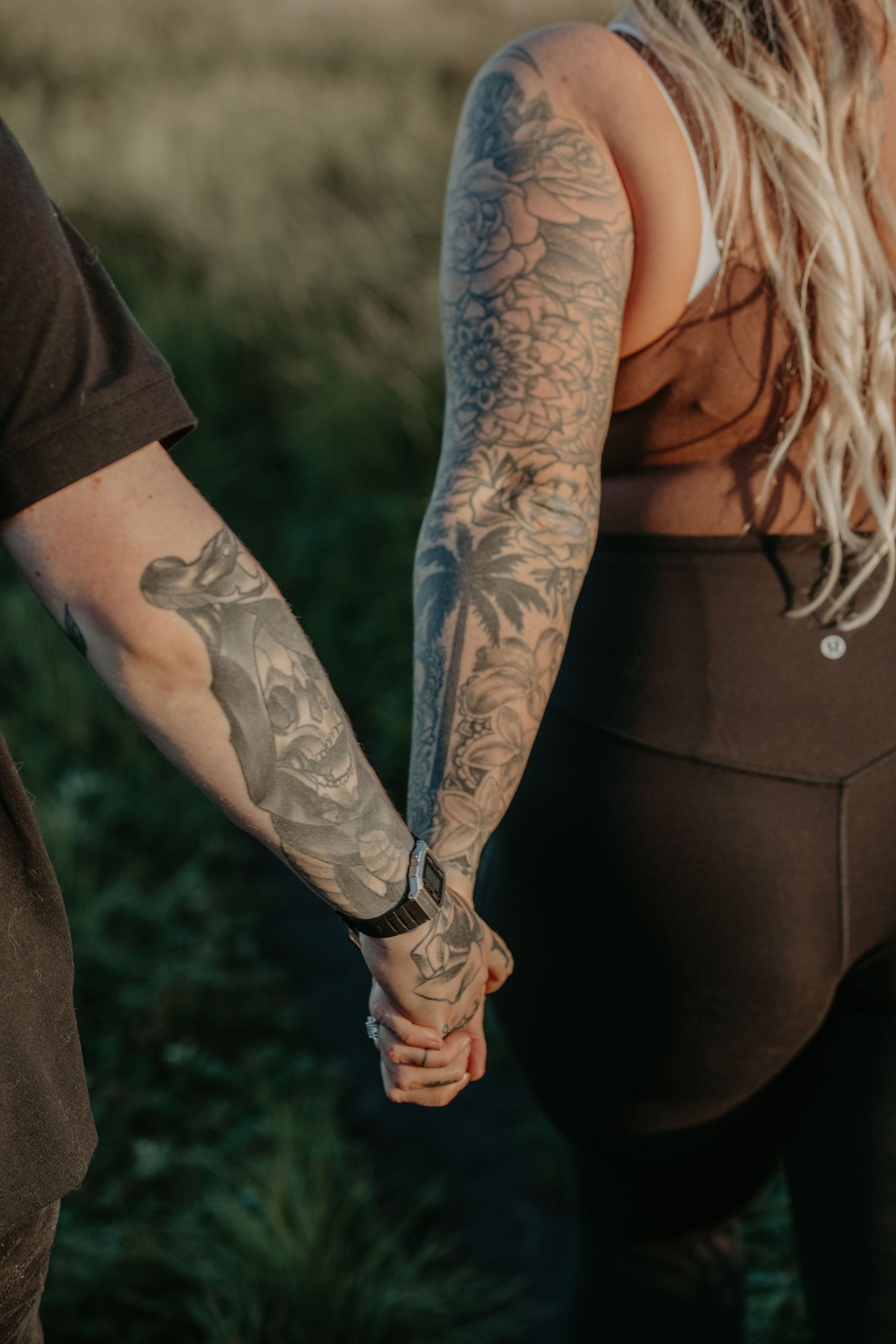 Close-up of tattooed couple holding hands outdoors with nature backdrop and brown clothing.