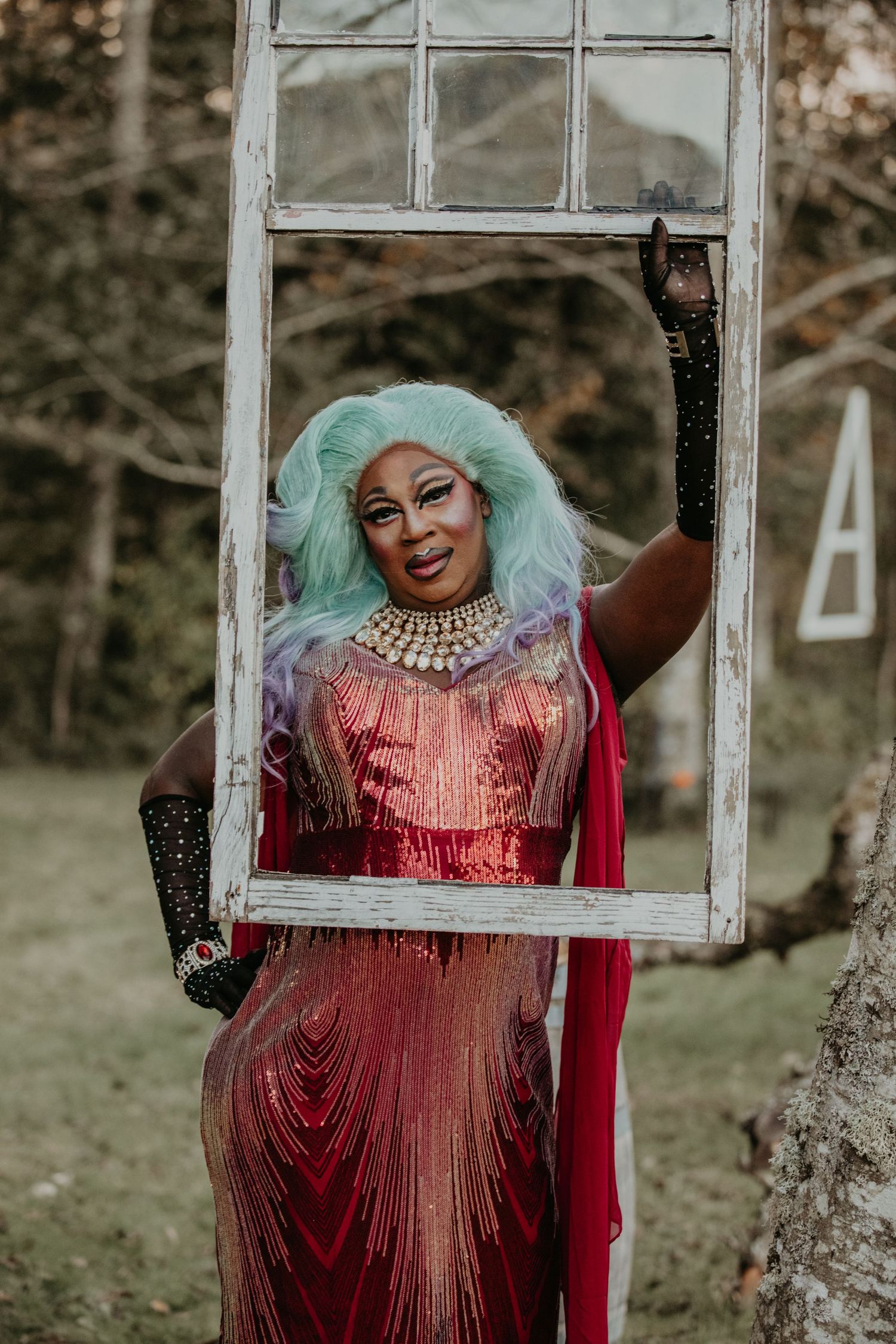 Person in green hair and red sequined gown poses behind vintage window frame during outdoor photoshoot.