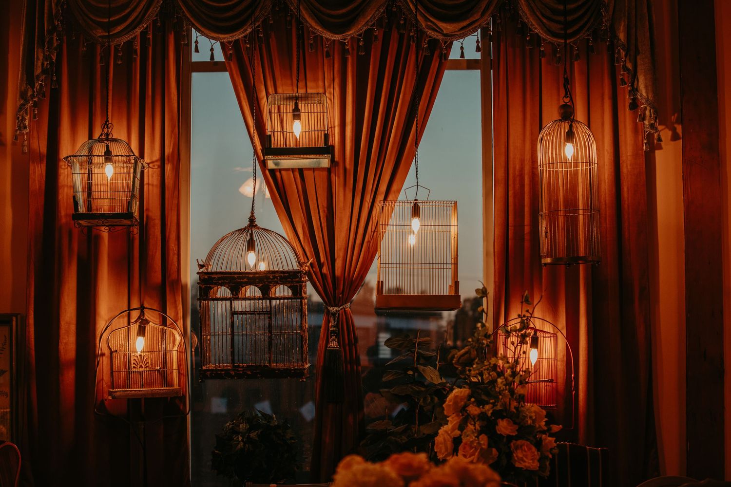 Vintage restaurant interior with red velvet curtains, glowing lanterns, and flowers silhouetted against window view at dusk.
