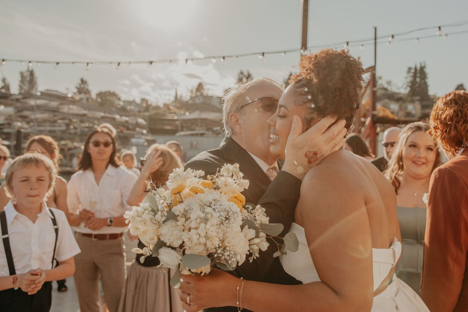 Wedding guests gather outdoors at sunset during a reception celebration with string lights and floral decorations.