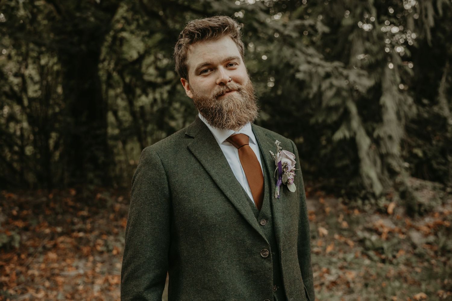 A wedding party poses in a rustic autumn forest setting wearing dark green suits with orange accents.