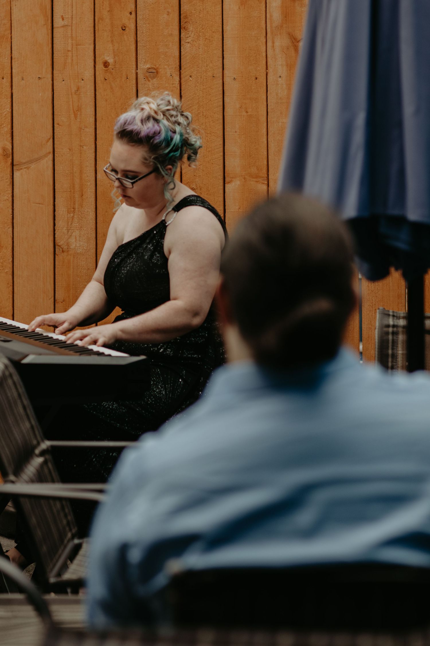 Musician in a black dress plays keyboard during performance with wooden paneled wall in background.
