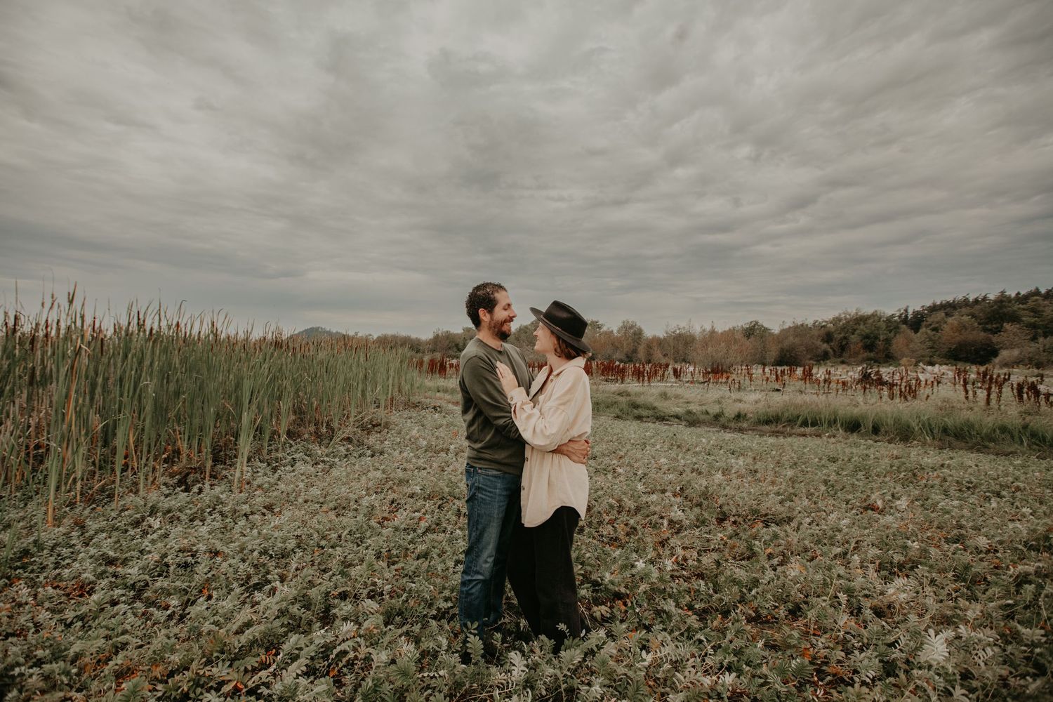 A couple embraces in a serene field with tall grasses under moody overcast skies on an autumn day.