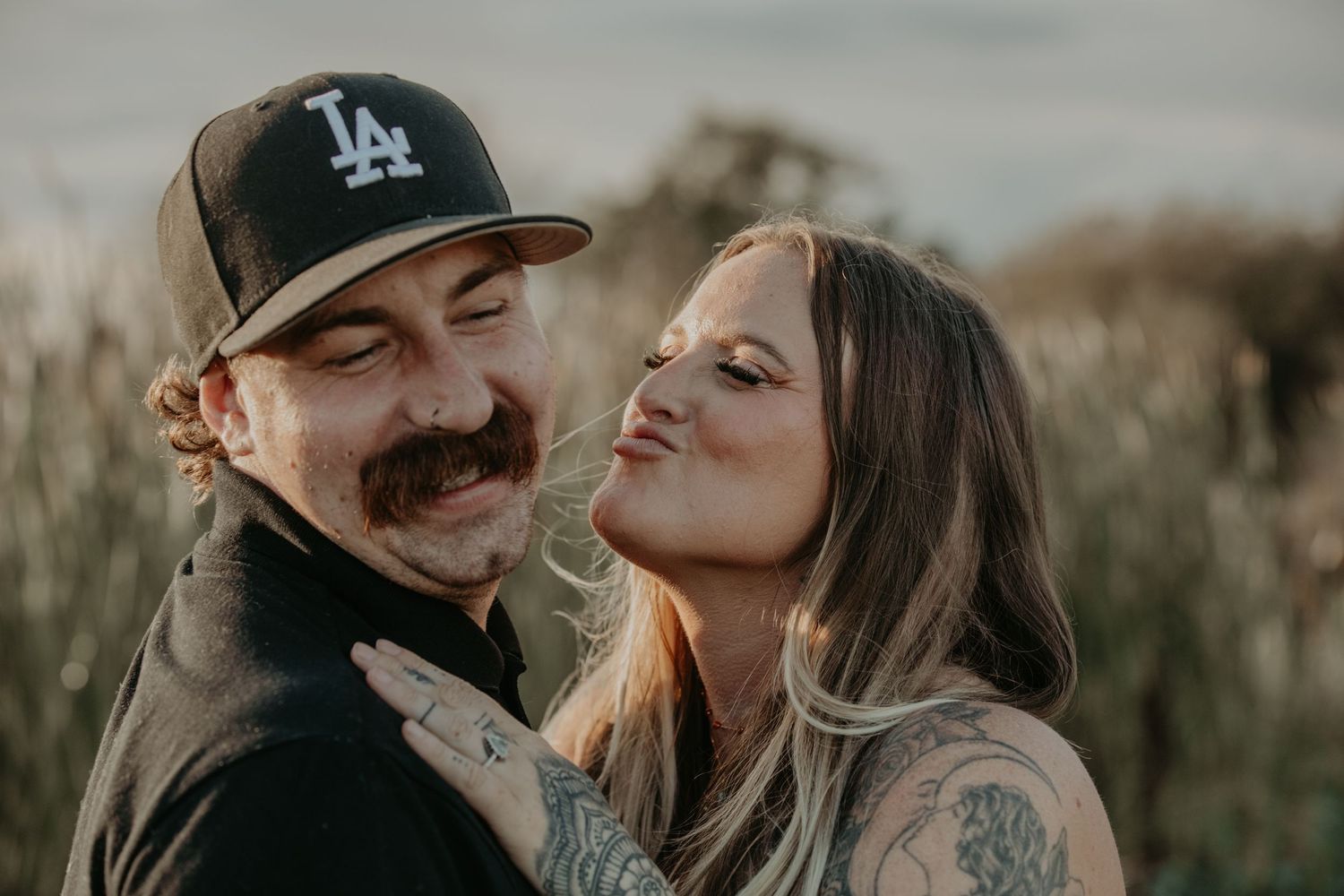 Couple shares intimate moment outdoors, wearing LA Dodgers hat against natural backdrop at golden hour.