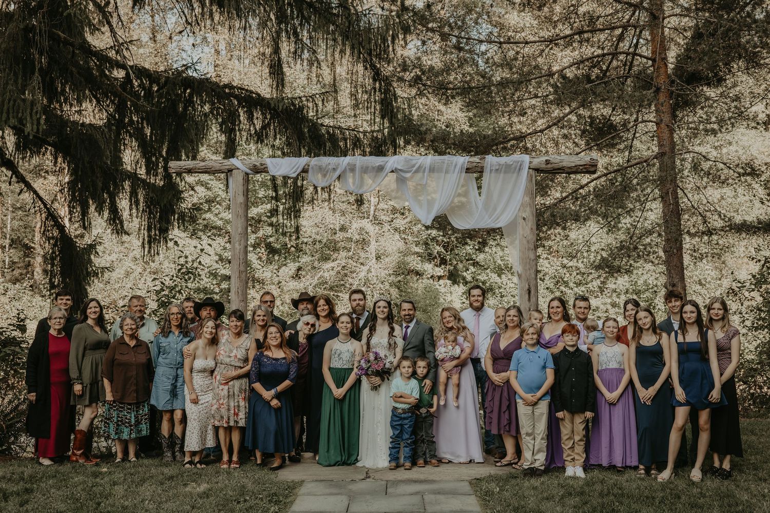 Large group gathered outdoors for a formal event wearing colorful dresses and suits against a rustic stone wall backdrop.