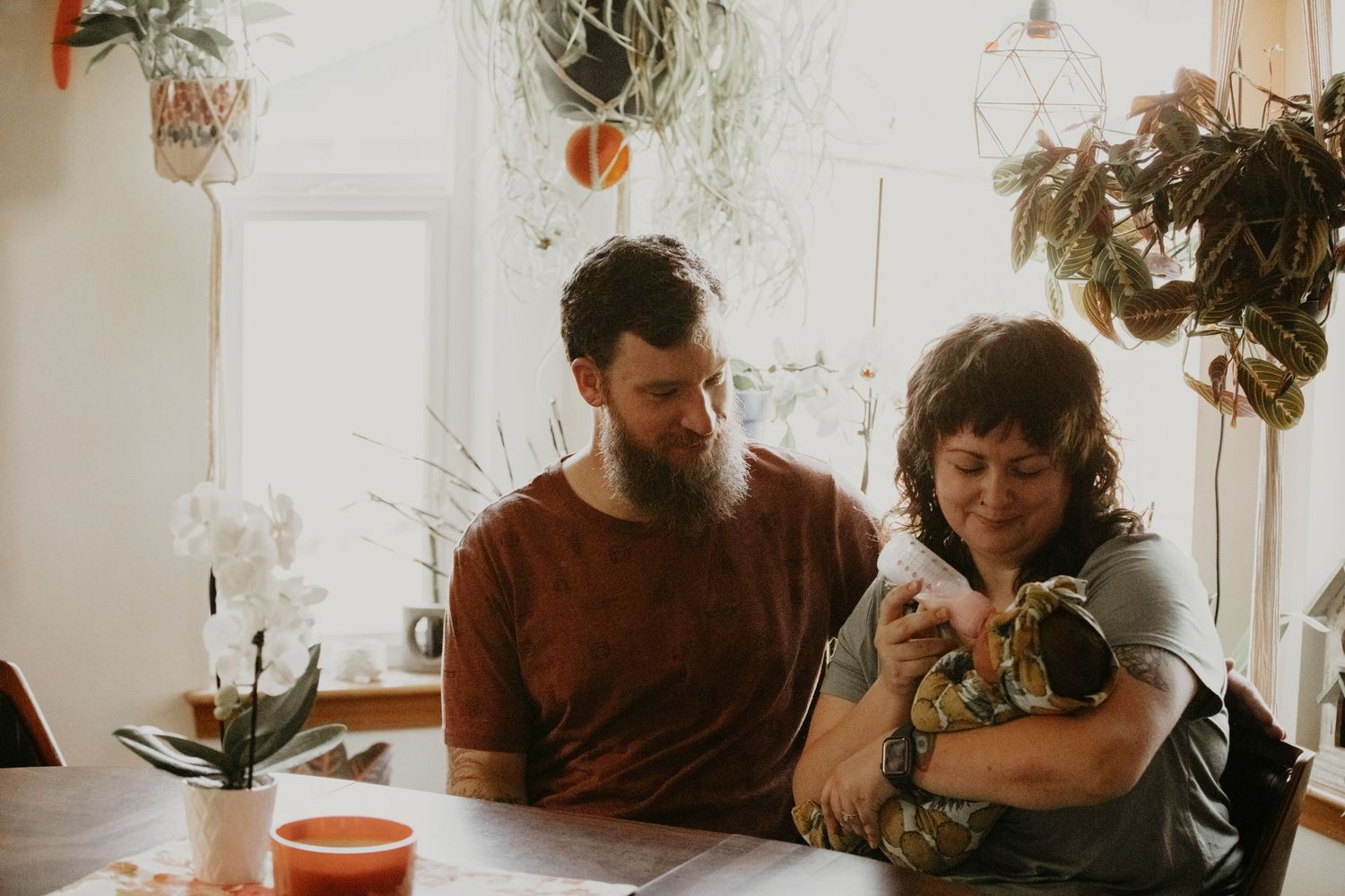 Two people sit at a table in a bright room filled with hanging plants while holding a small pet.