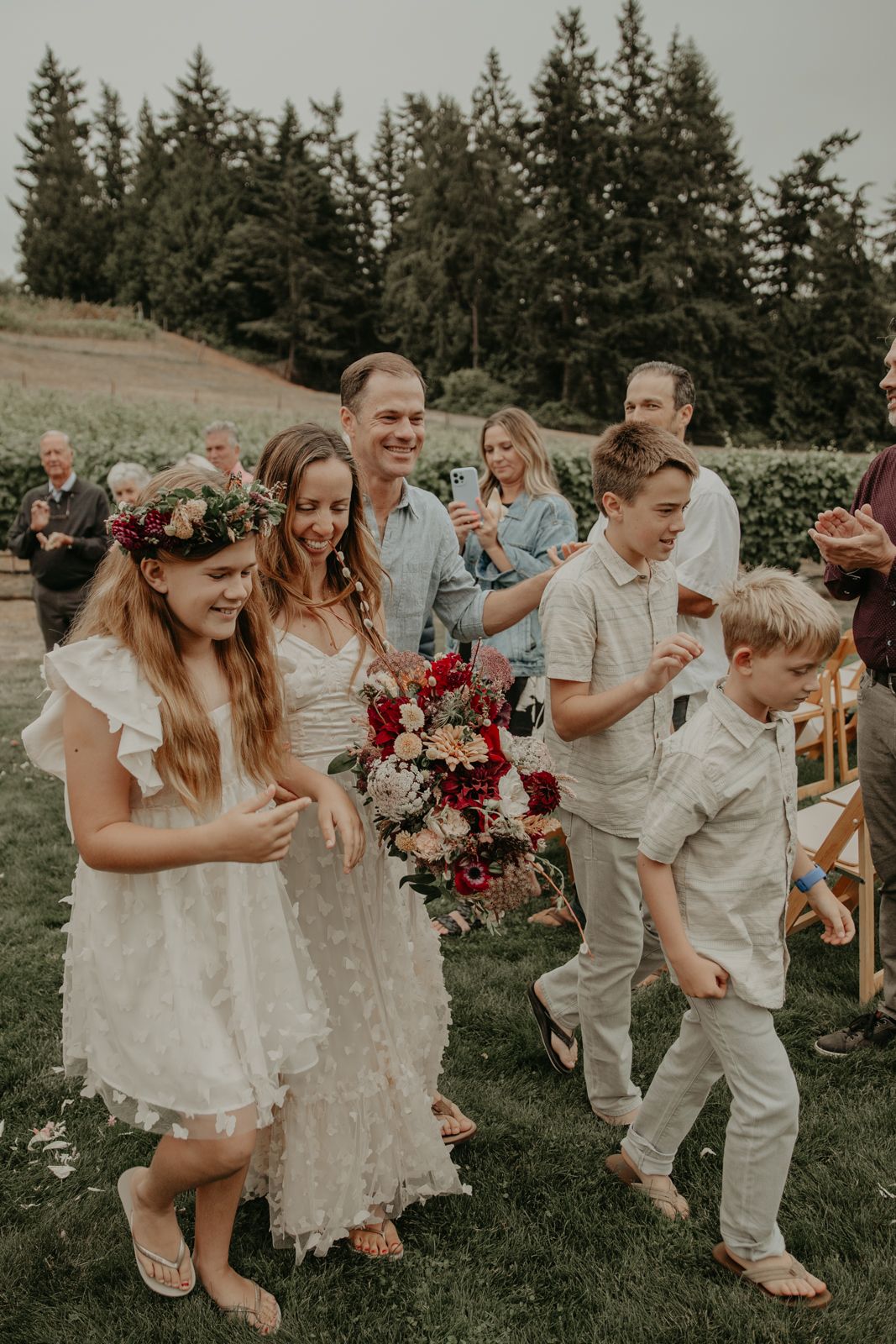 Wedding party dances and celebrates on a grassy outdoor venue with evergreen trees in the background.