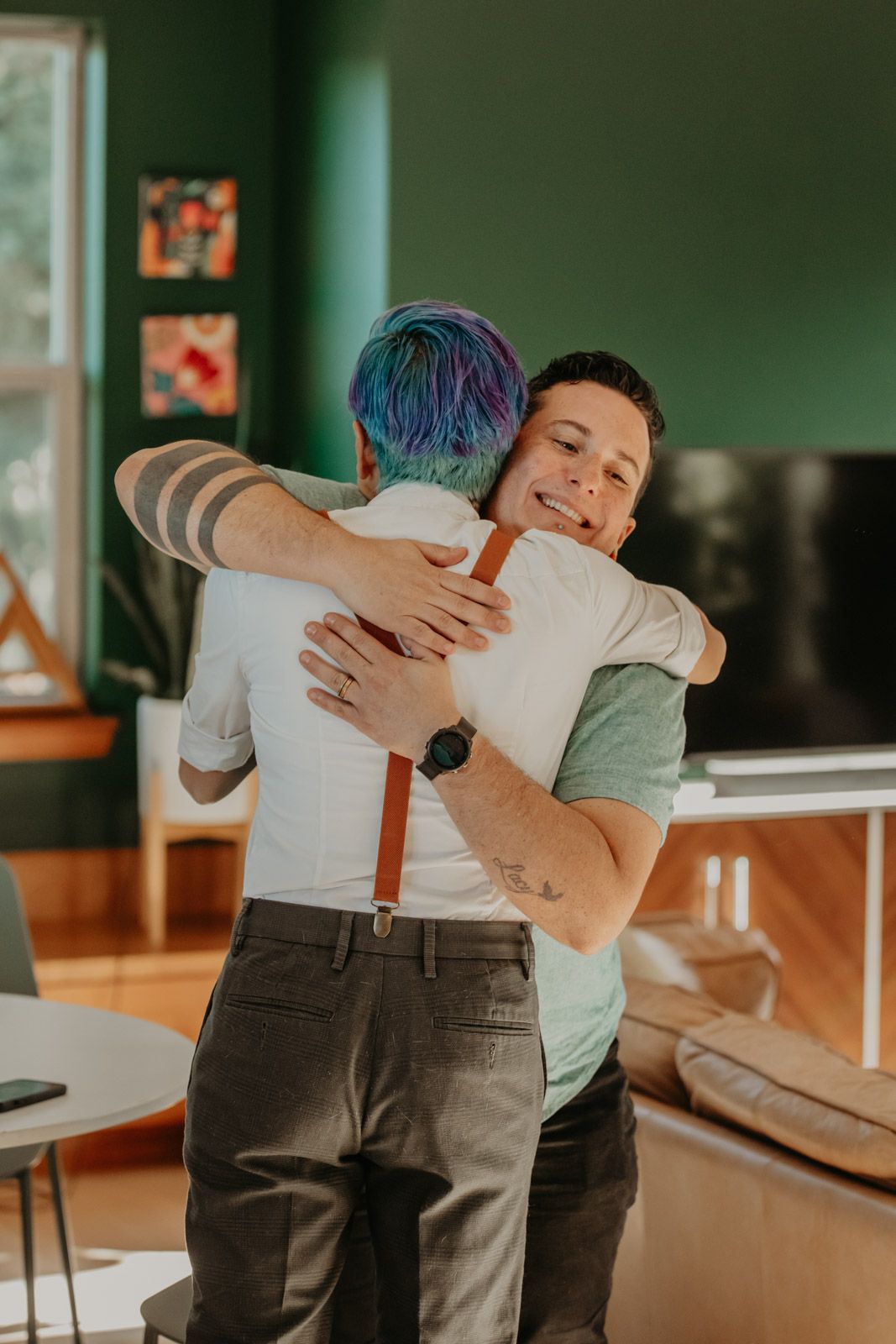 Two people share a warm embrace in an indoor setting with green walls and natural lighting.
