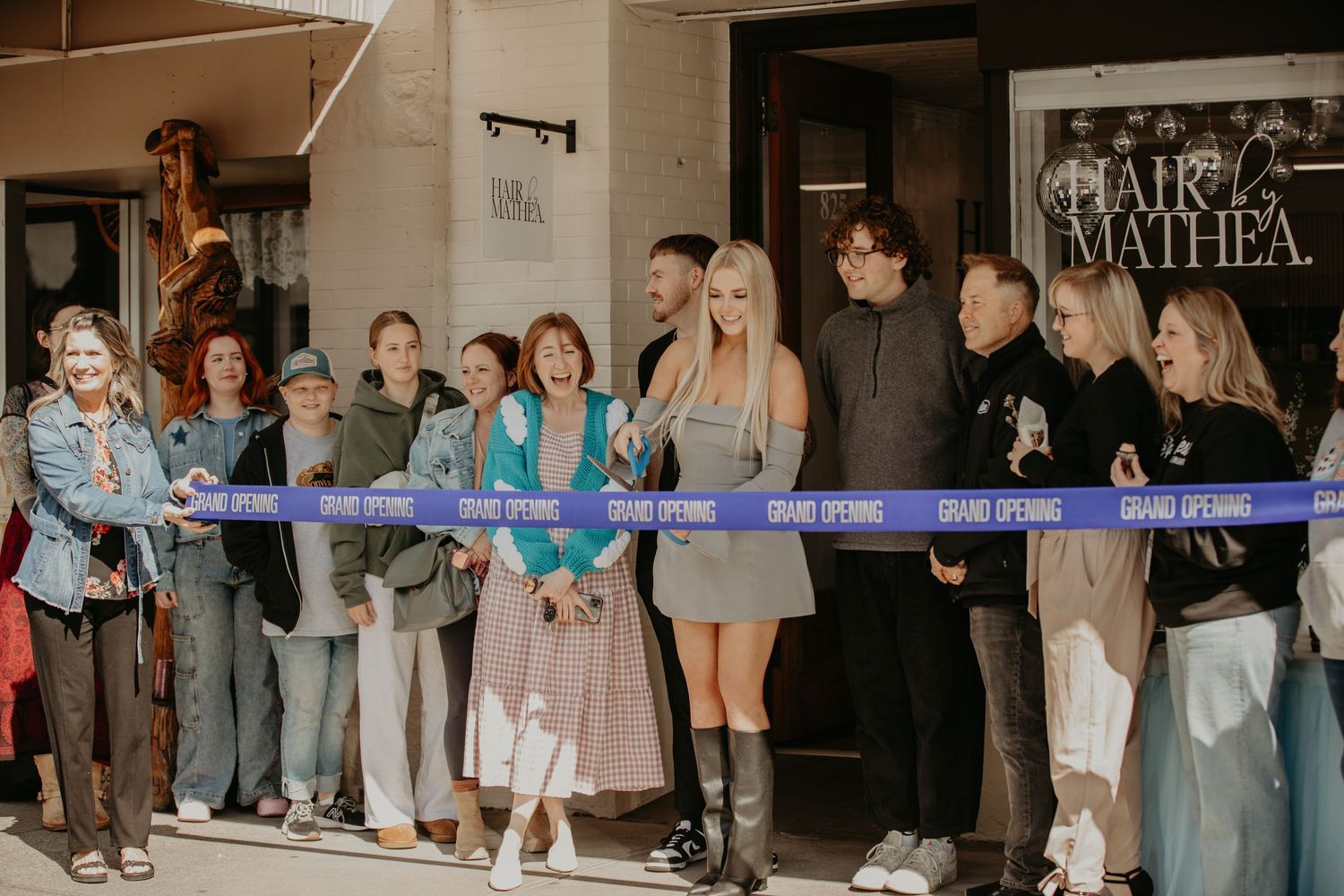 A large group of people stand together at a ribbon cutting ceremony outside a business entrance.