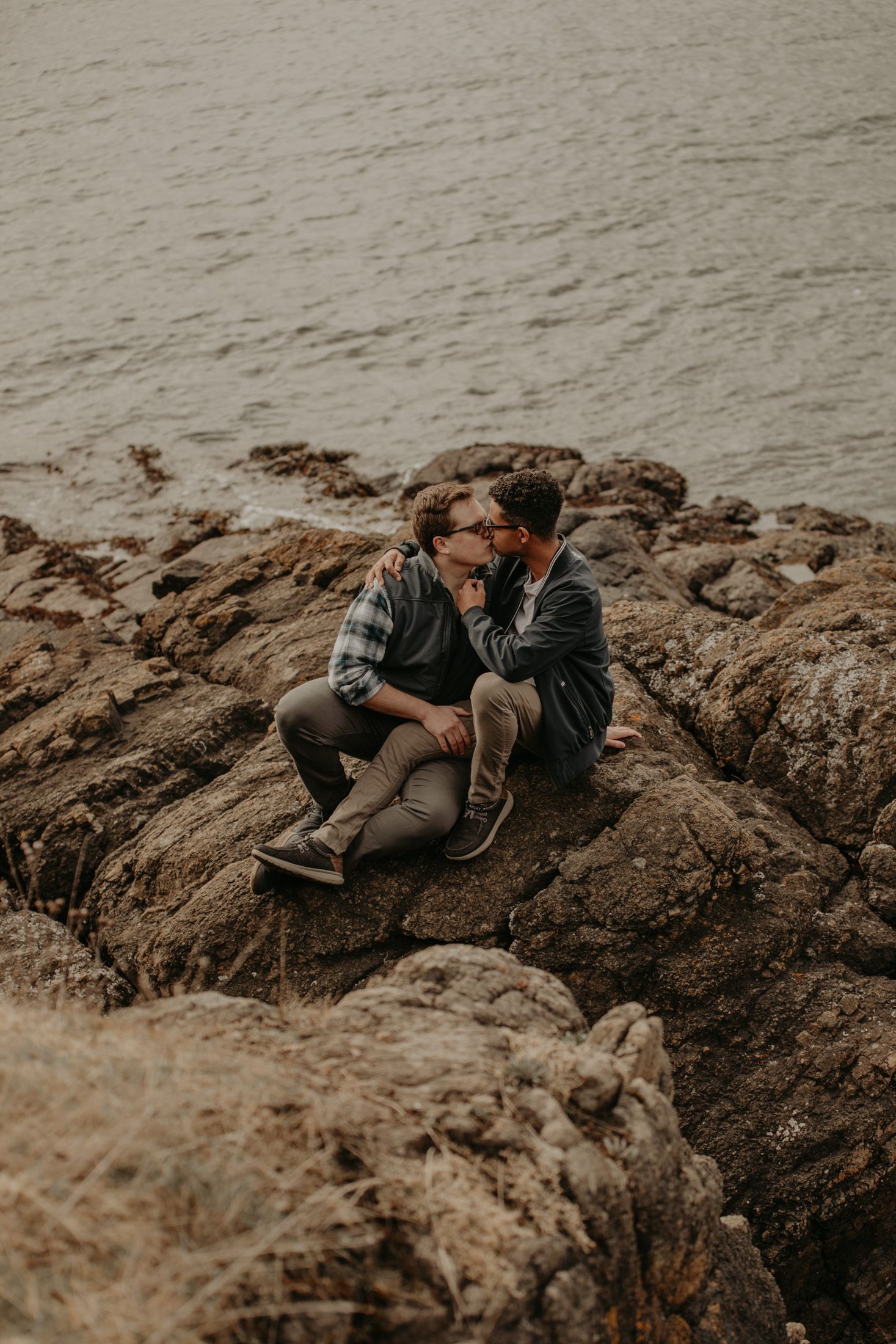 Two people share a romantic moment sitting on rocky coastal cliffs with calm ocean waters in the background.