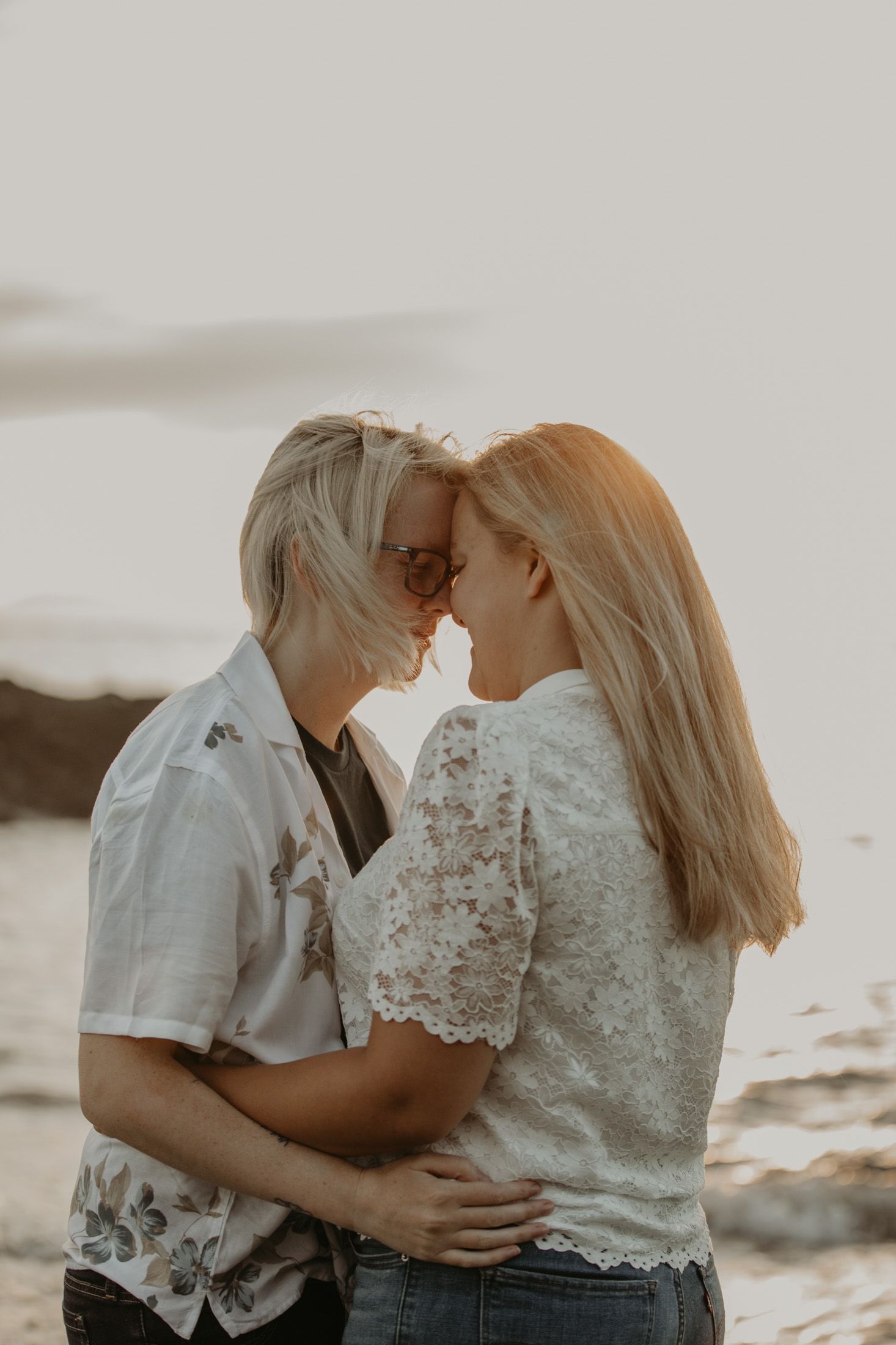 Two blonde women share a romantic forehead touch on a beach at sunset, wearing white tops and embracing each other.